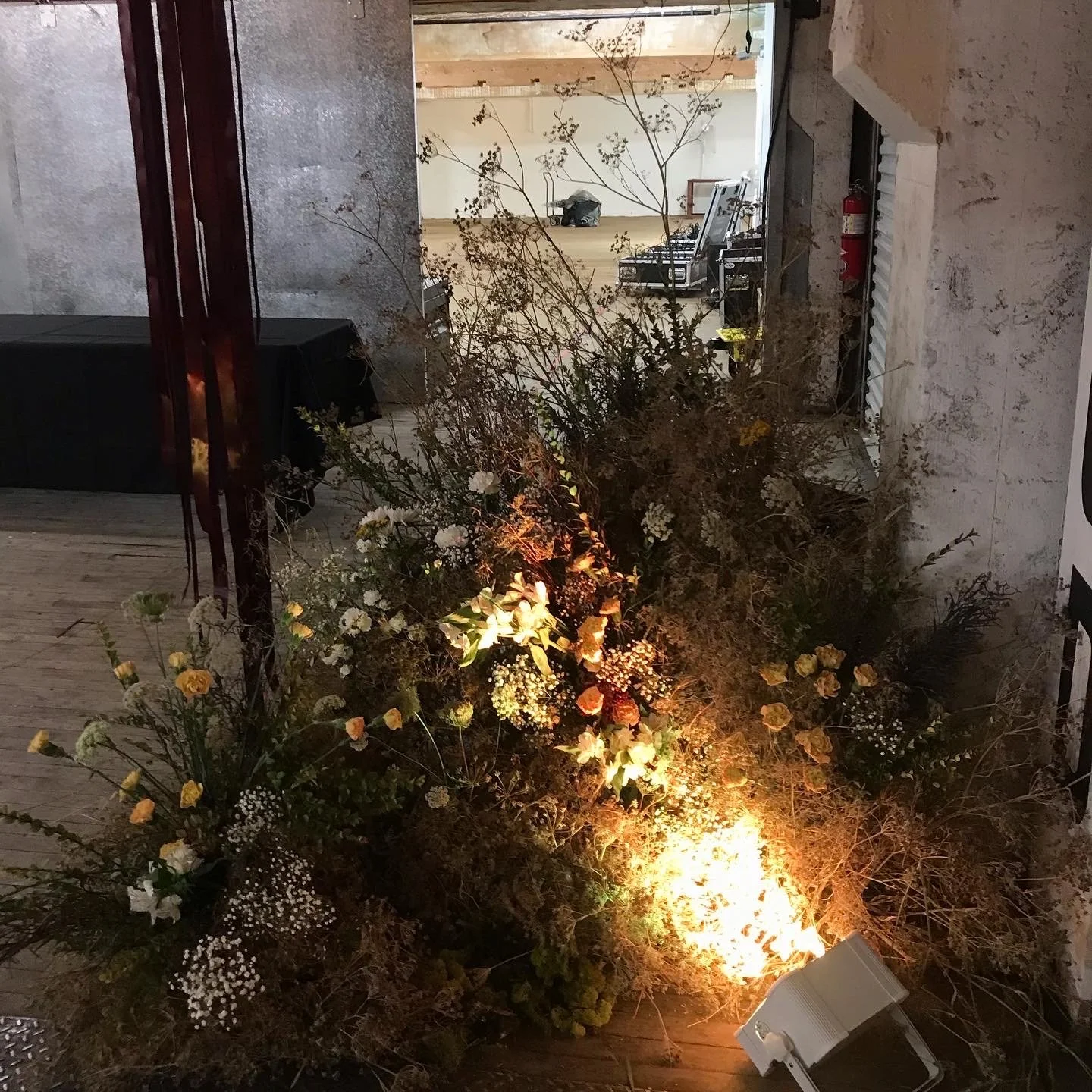 A large bouquet of mixed flowers illuminated by a small warm-colored light, situated near a textured wall and adjacent to a doorway, with a view of an open space with equipment and furniture in the background.