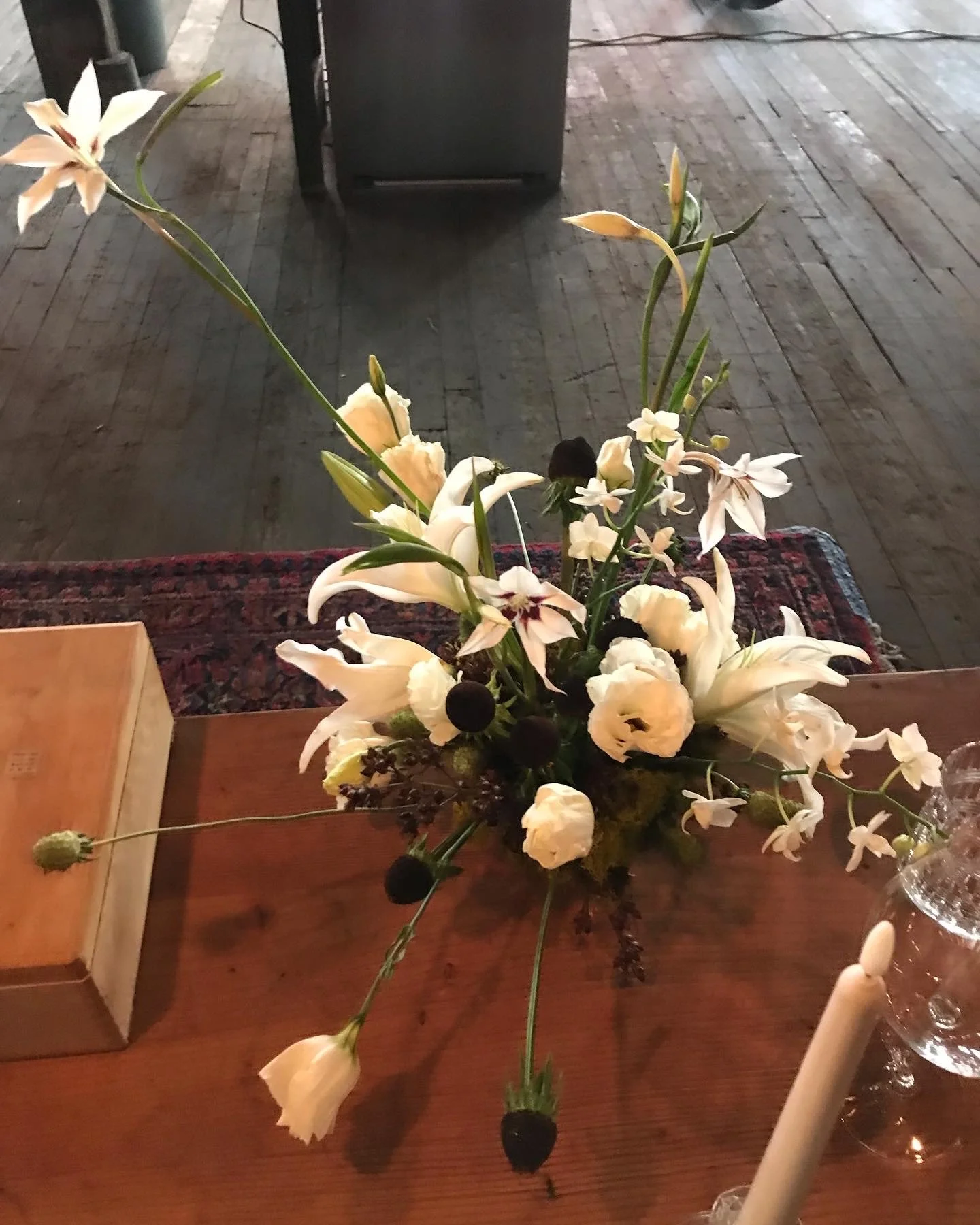 A bouquet of white flowers on a wooden table, with a candle and glassware nearby, in a dimly lit setting.
