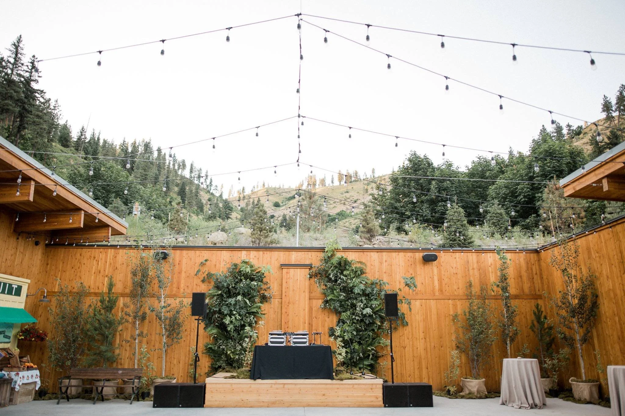 Outdoor stage with string lights, trees, wooden fencing, and mountain scenery in the background, set up for a performance or event.