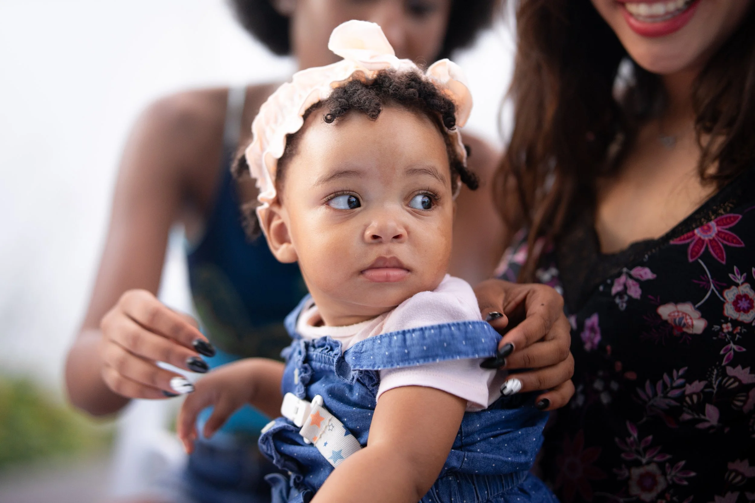 Close-up of a young girl with curly hair wearing a pink headband, sitting on a woman's lap, looking sideways with a curious expression. The woman is smiling, and another person in the background is touching her shoulder.