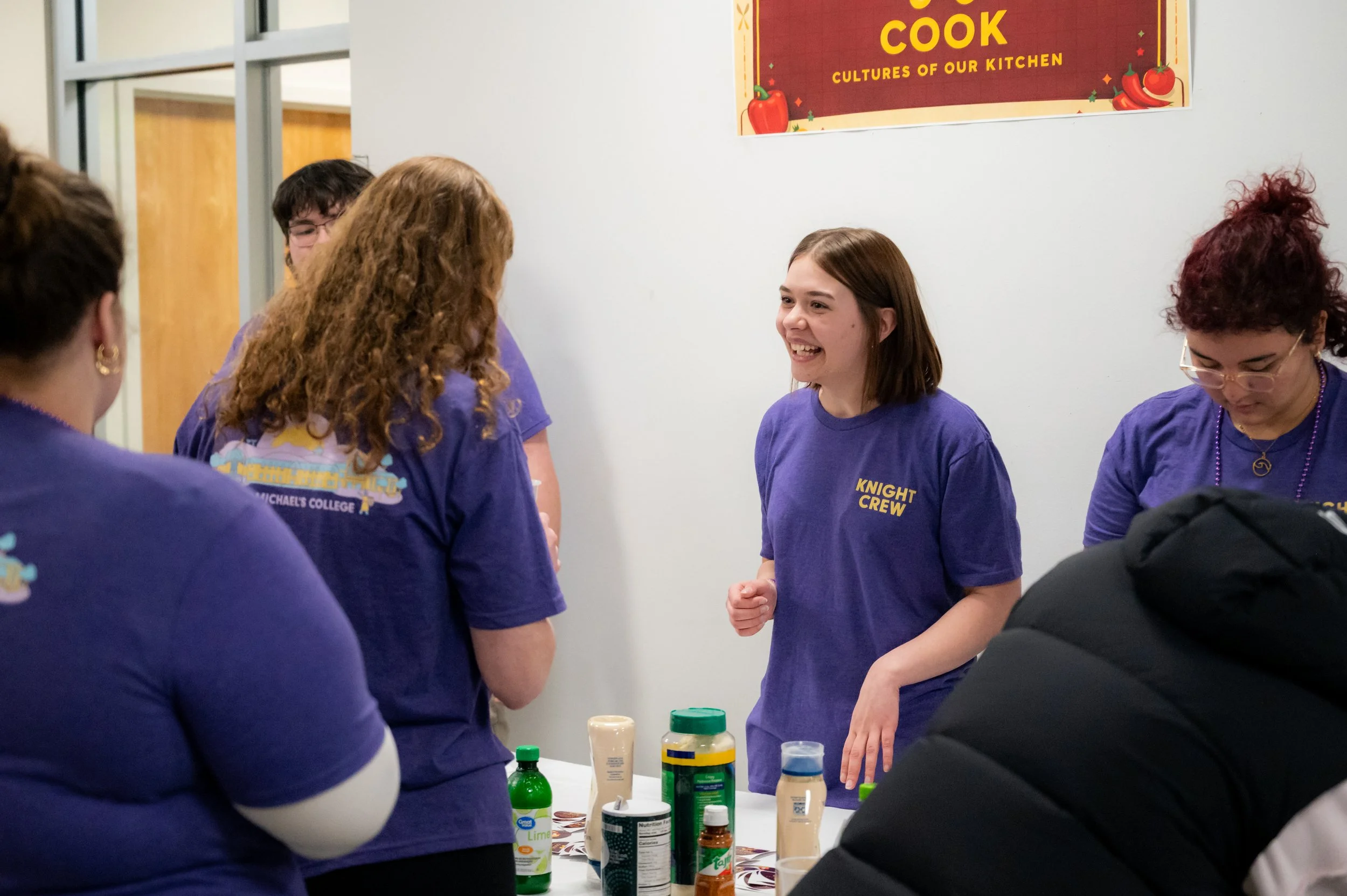 Group of young women wearing purple shirts, with one smiling and talking, standing around a table with food and condiments, in a room with a sign that says 'COOK CULTURES OF OUR KITCHEN'