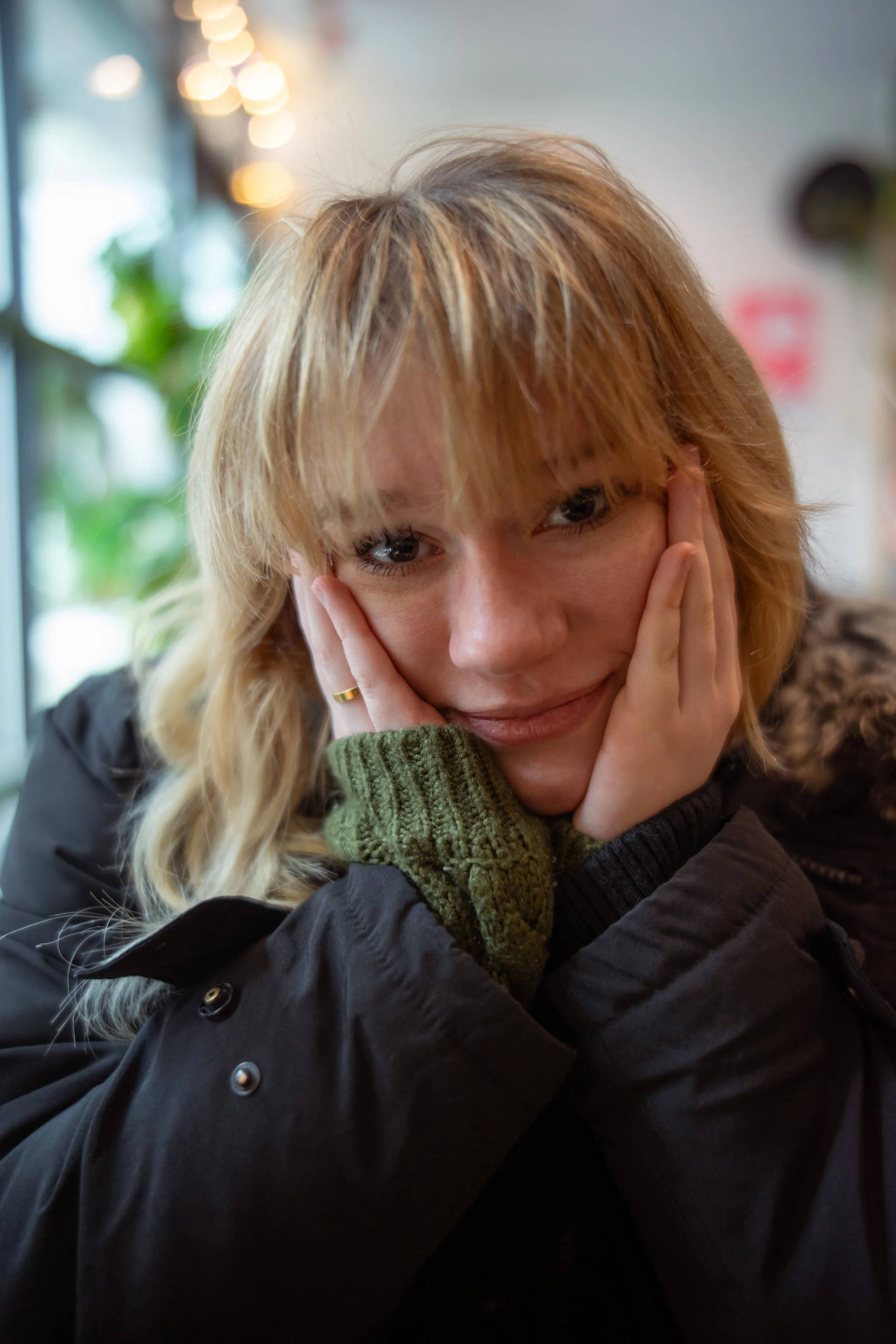 A woman with blonde hair and wearing a black jacket and green knit sweater rests her face in her hands in a cozy indoor setting with warm lighting and blurred background.