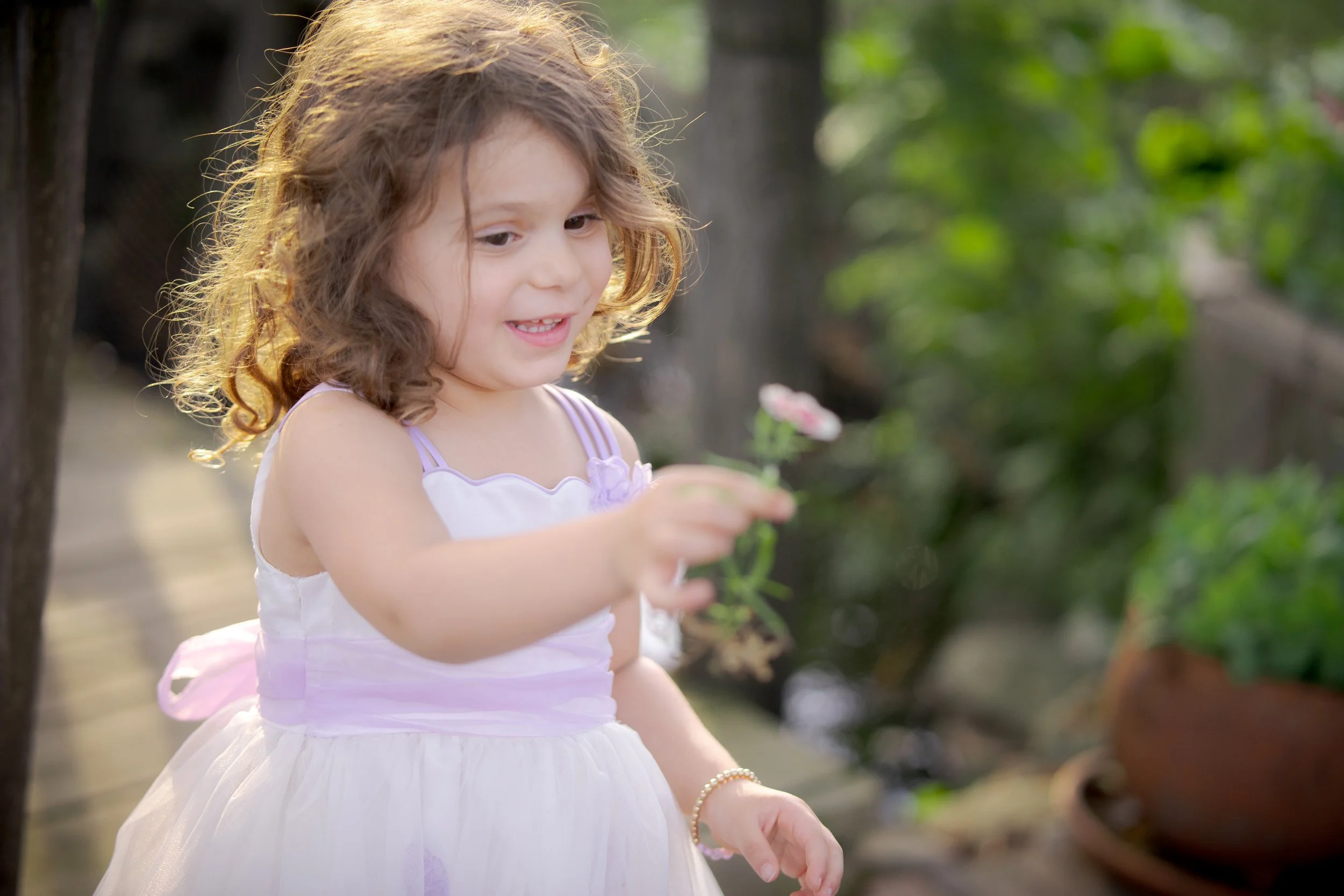 A young girl with curly brown hair and a white dress with purple accents is holding a flower and smiling outdoors with green foliage in the background.