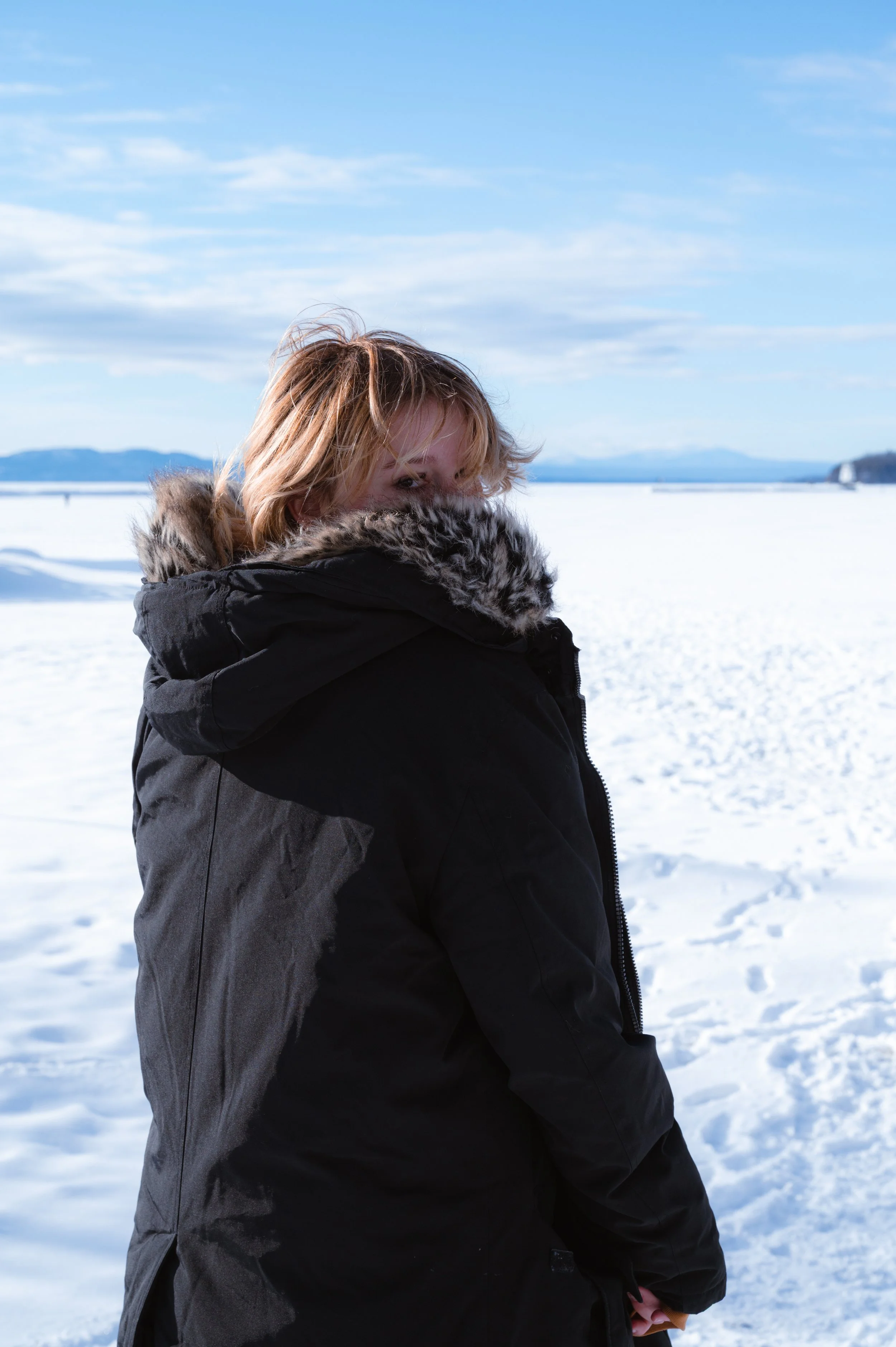 A woman with light brown hair, partially covered by a fur-lined hood, standing in a snowy landscape with distant mountains and a blue sky.