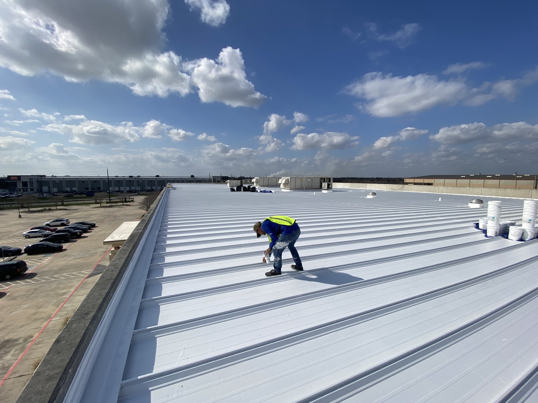 A worker on a rooftop installing white roofing material amidst a clear blue sky with scattered clouds. There are parked cars and a large commercial building in the background.