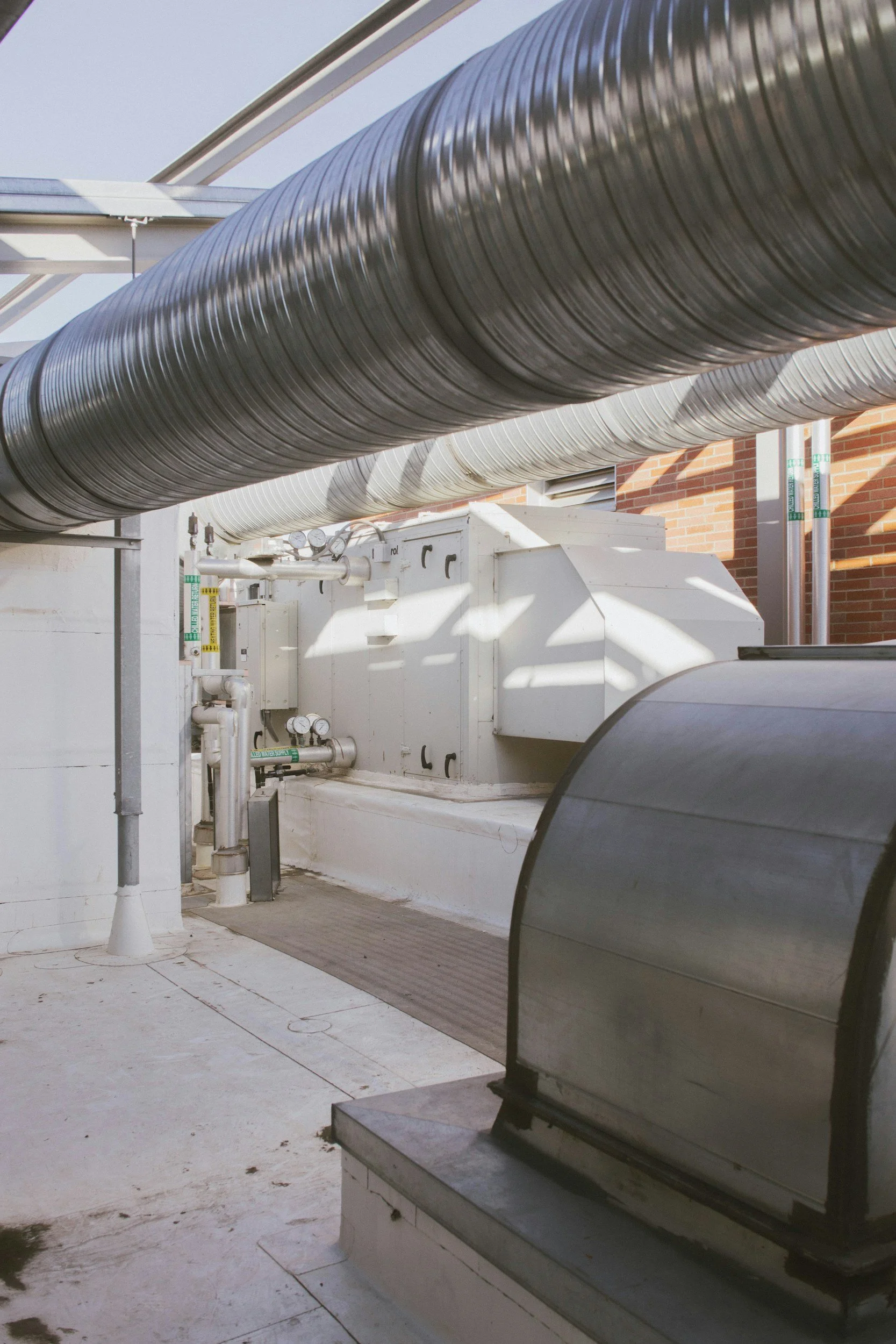 A rooftop area with large metallic ventilation pipes running parallel to each other, a brick building wall in the background, and an ice machine or similar equipment in the foreground.