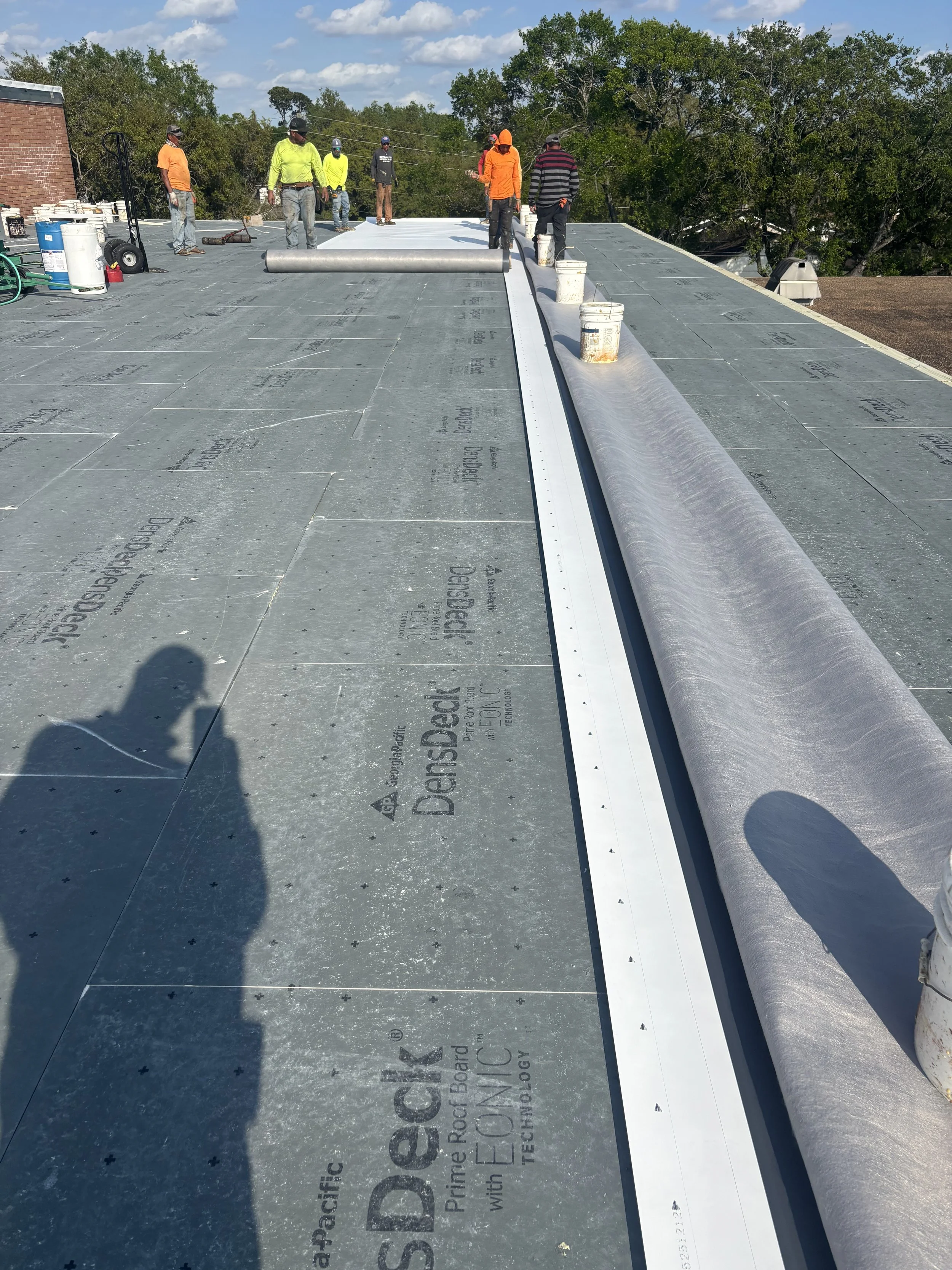 Construction workers installing roofing material on a flat roof, with tools and buckets nearby, surrounded by trees and under a partly cloudy sky.