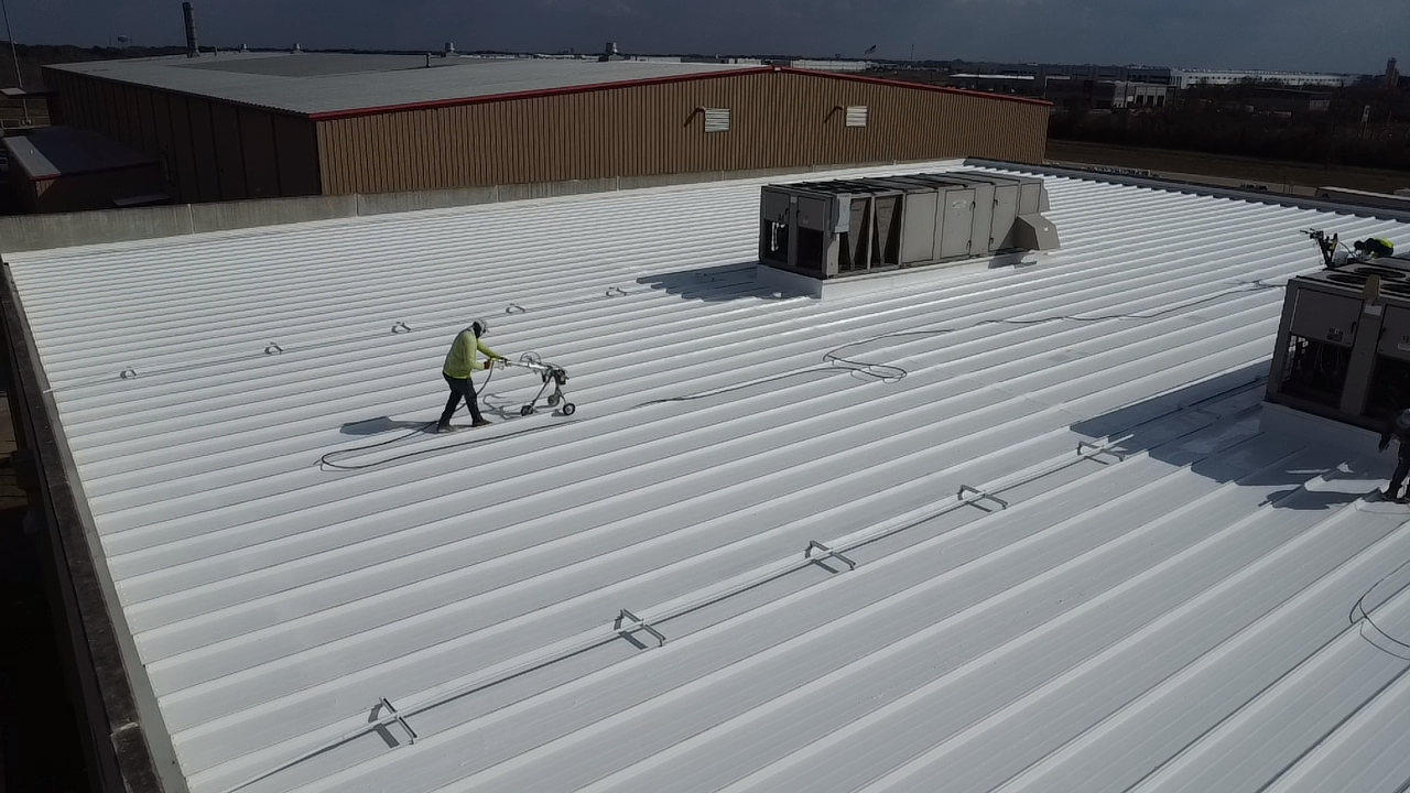 Workers cleaning a large white metal roof with cleaning equipment, with ventilation units and a brown building in the background.