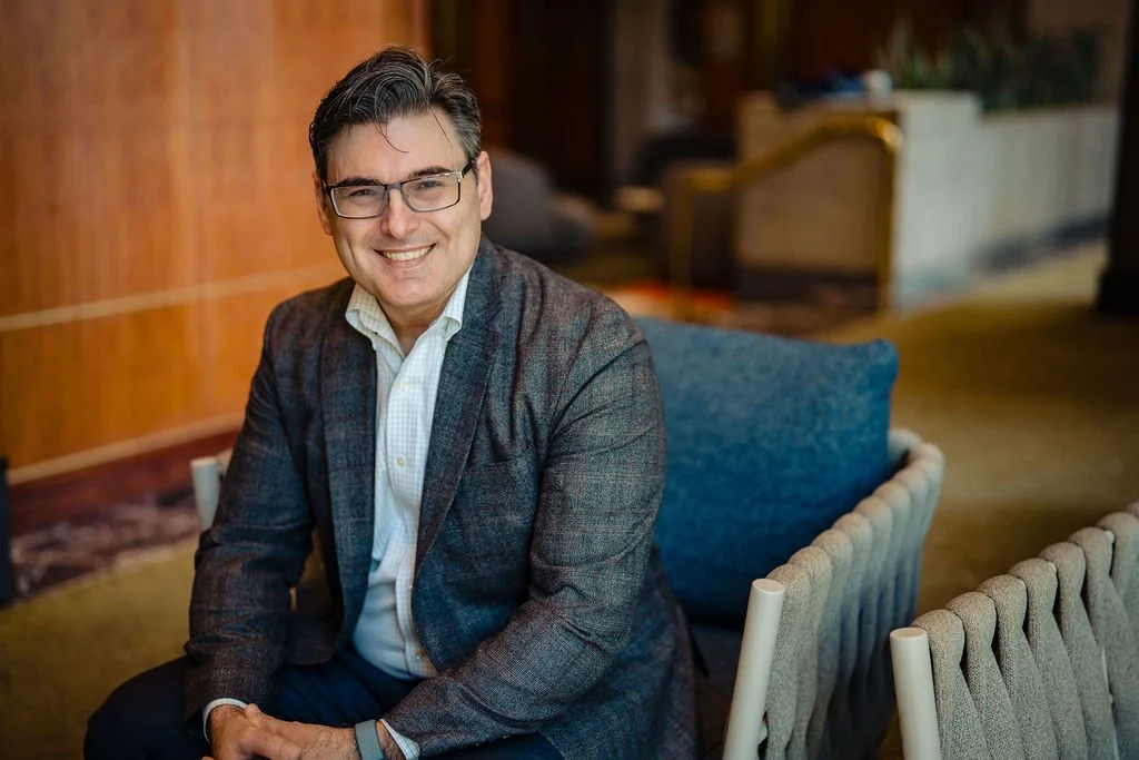 A smiling man with glasses and dark hair in a plaid blazer, sitting on a blue cushion in a warmly lit room.