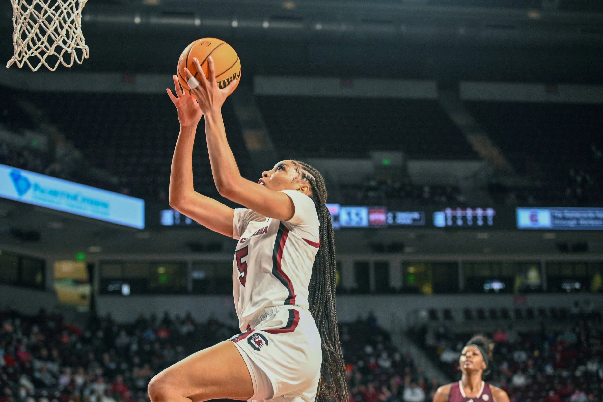 A female basketball player in a white and maroon uniform jumps to shoot the ball towards the hoop inside an indoor basketball arena.