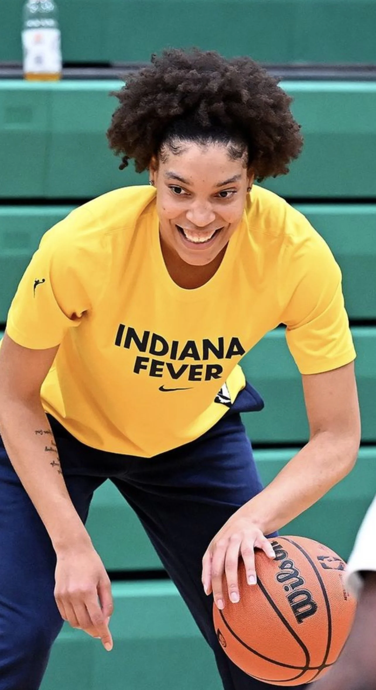 A woman with curly hair wearing a yellow Indiana Fever t-shirt dribbling a basketball on an indoor court.