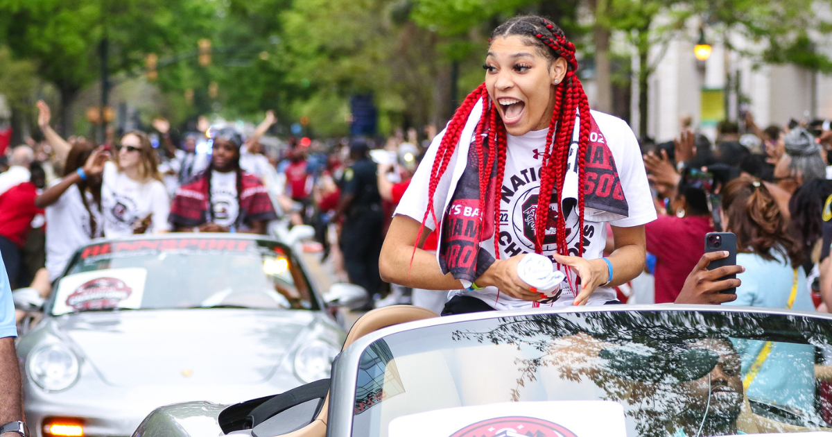 A young woman with red braids rides in a convertible car at a parade, smiling and holding a rolled-up poster, surrounded by a crowd of spectators and participants celebrating.