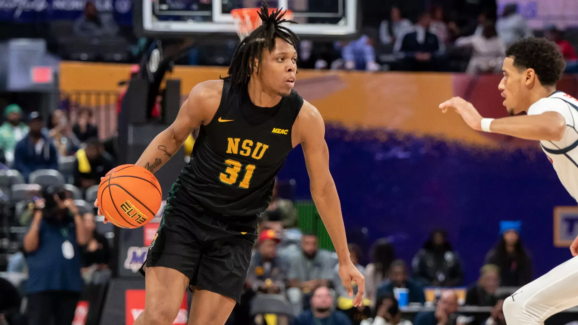 A basketball player in a black NSU uniform dribbling the ball and advancing past a defender in a white uniform during a game.