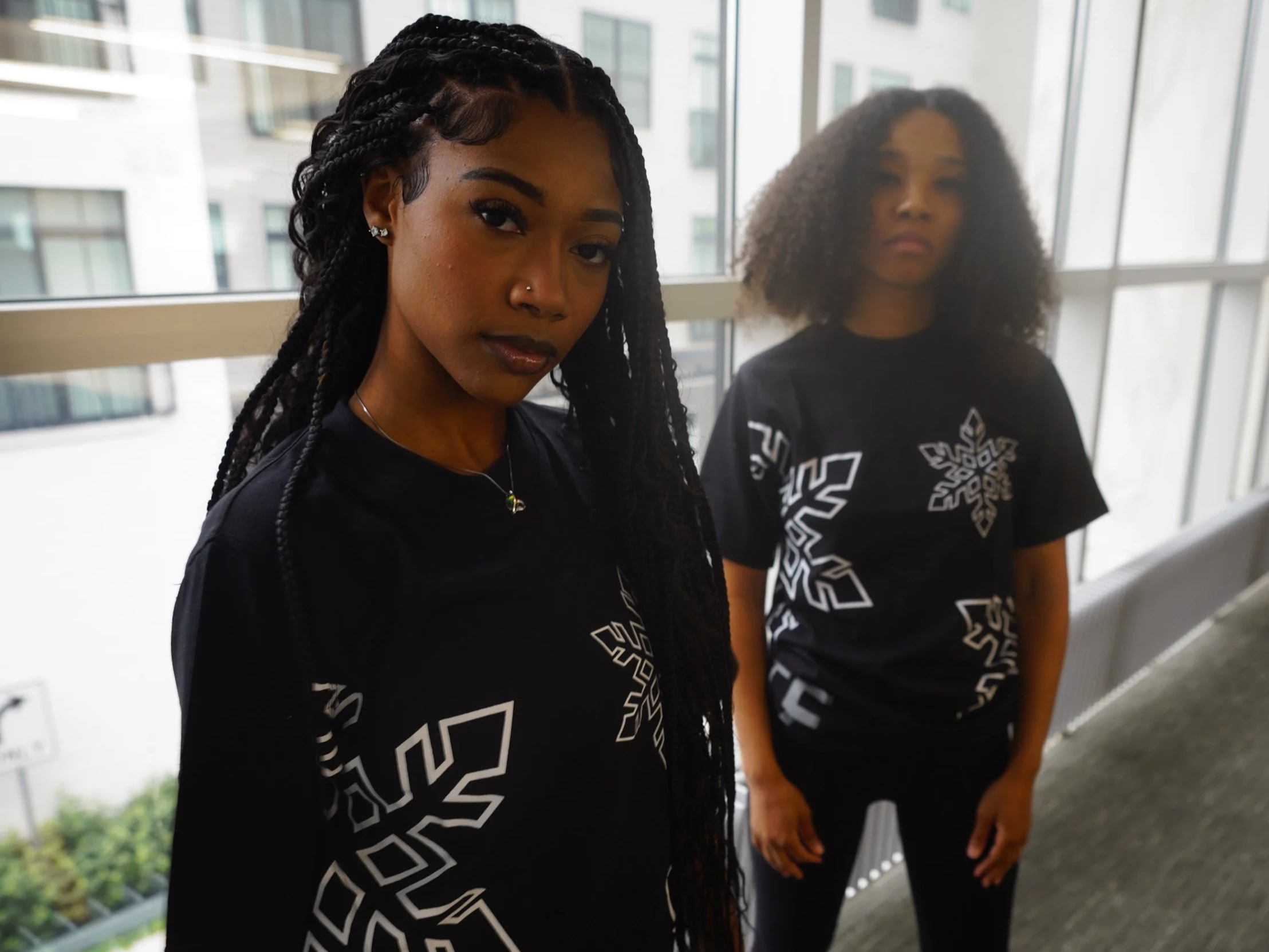 Two women standing side by side indoors, wearing black shirts with white graphic designs, near large windows showing a cityscape with tall buildings.