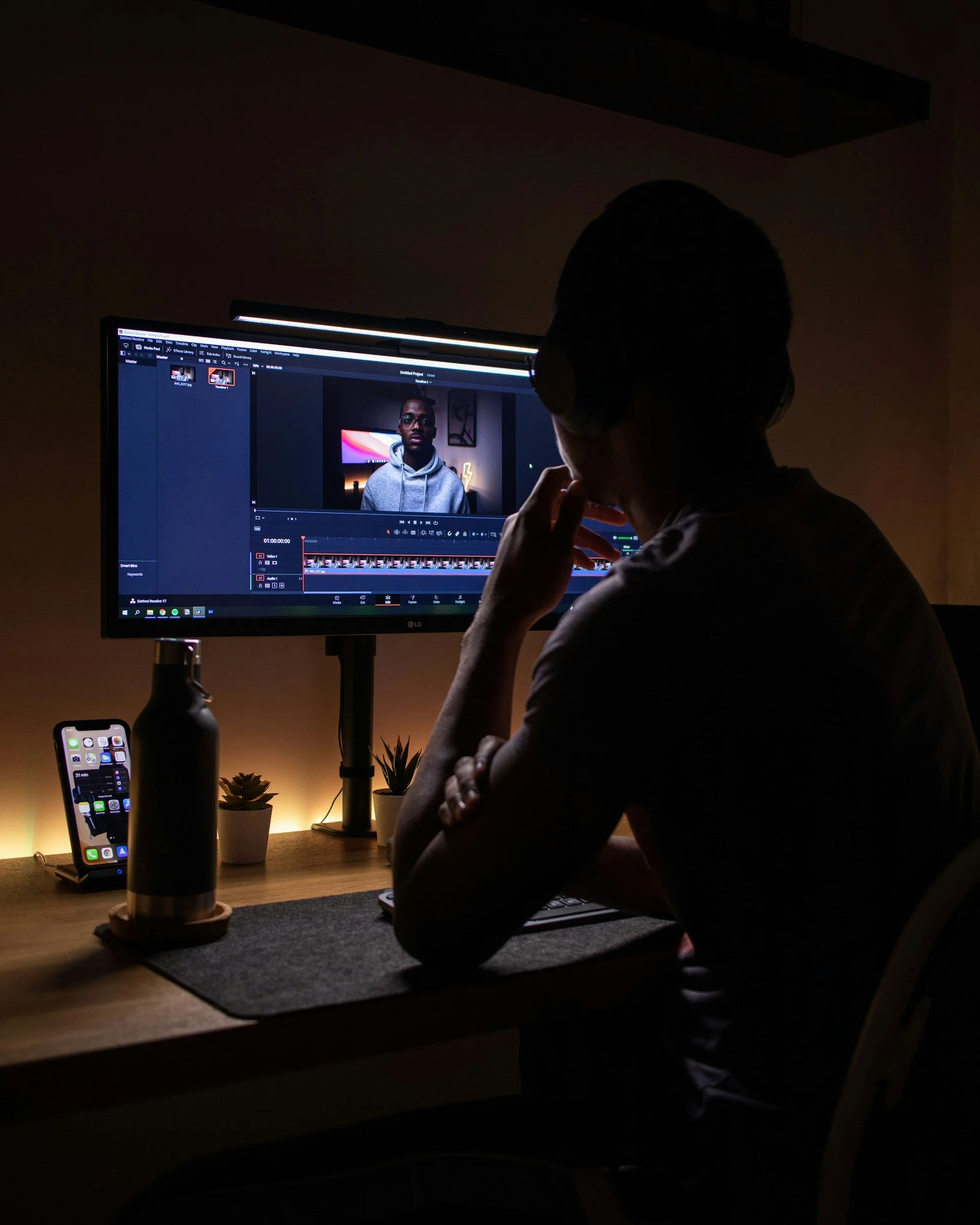 A person working on video editing at a desk with dim lighting. The computer screen shows a video editing software interface, and there are two small potted plants and a smartphone on the desk. The person is wearing headphones and appears focused.