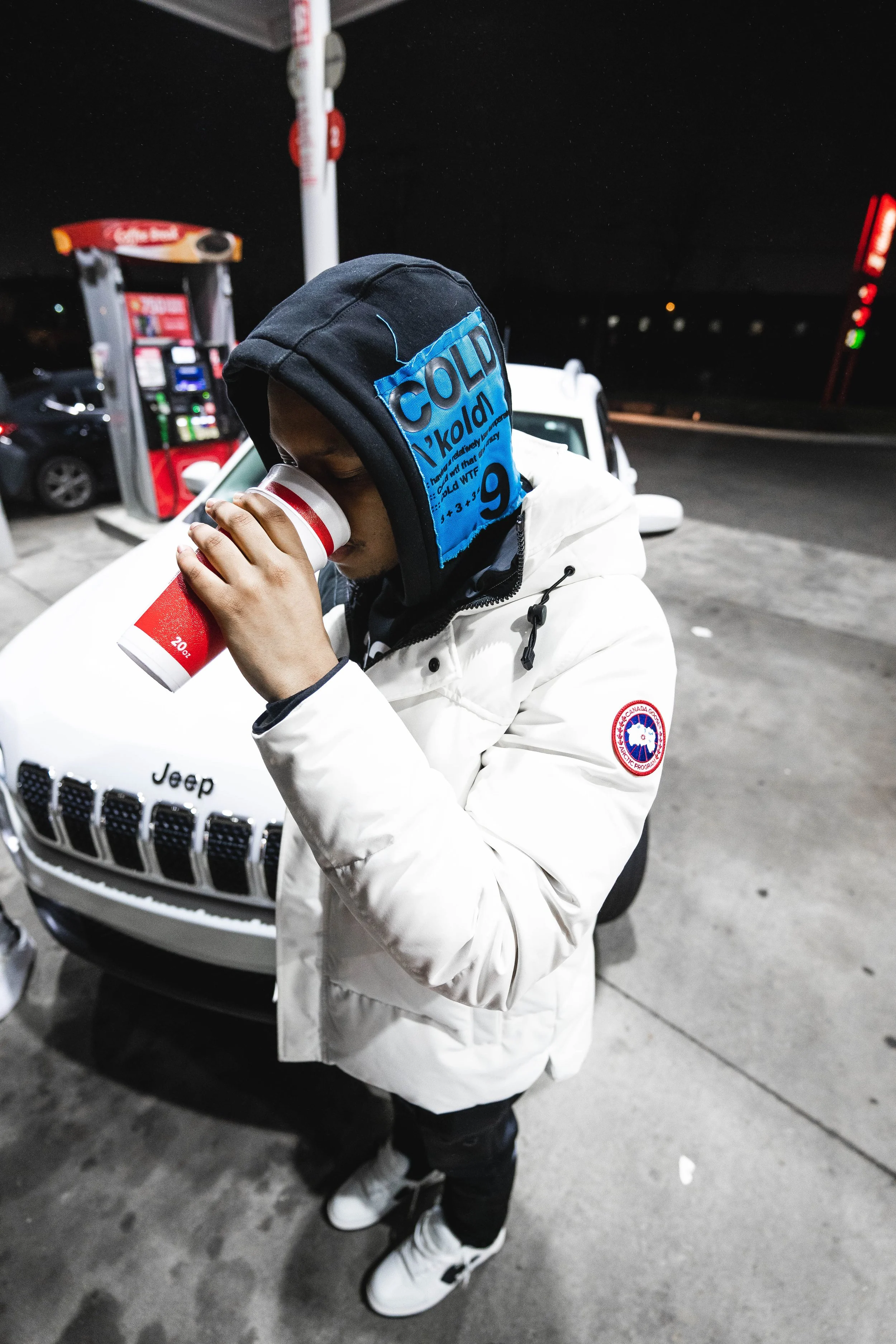 A person wearing a black hoodie with blue text and a white Canada Goose jacket drinking coffee at a gas station.