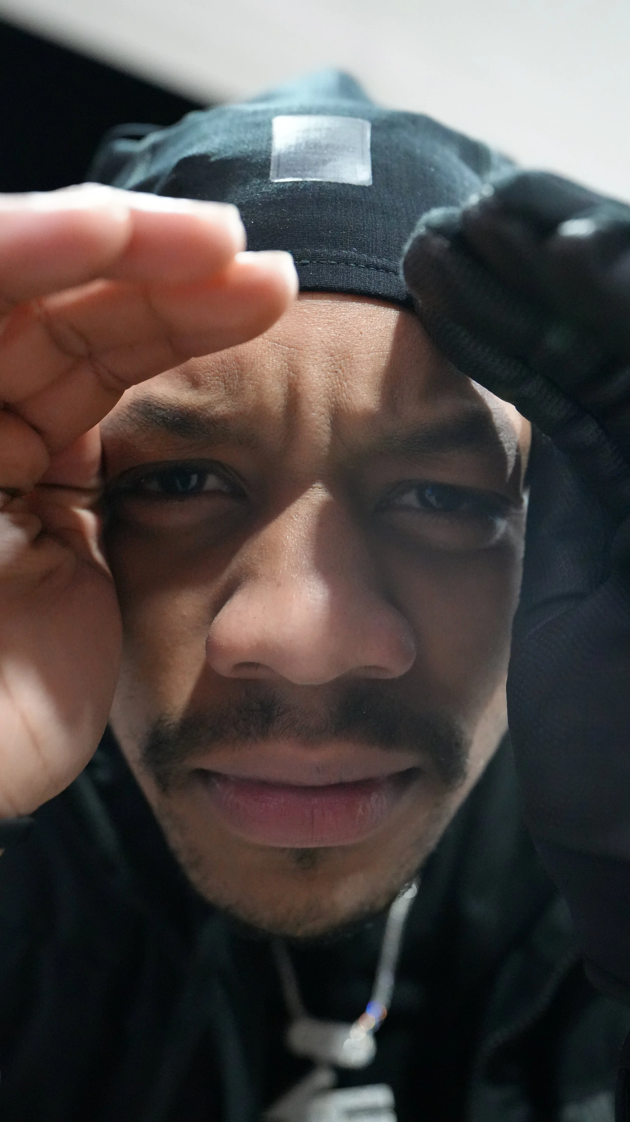 Close-up of a man with a mustache and beard, wearing a black beanie and black jacket, holding his right hand near his eye and smiling.