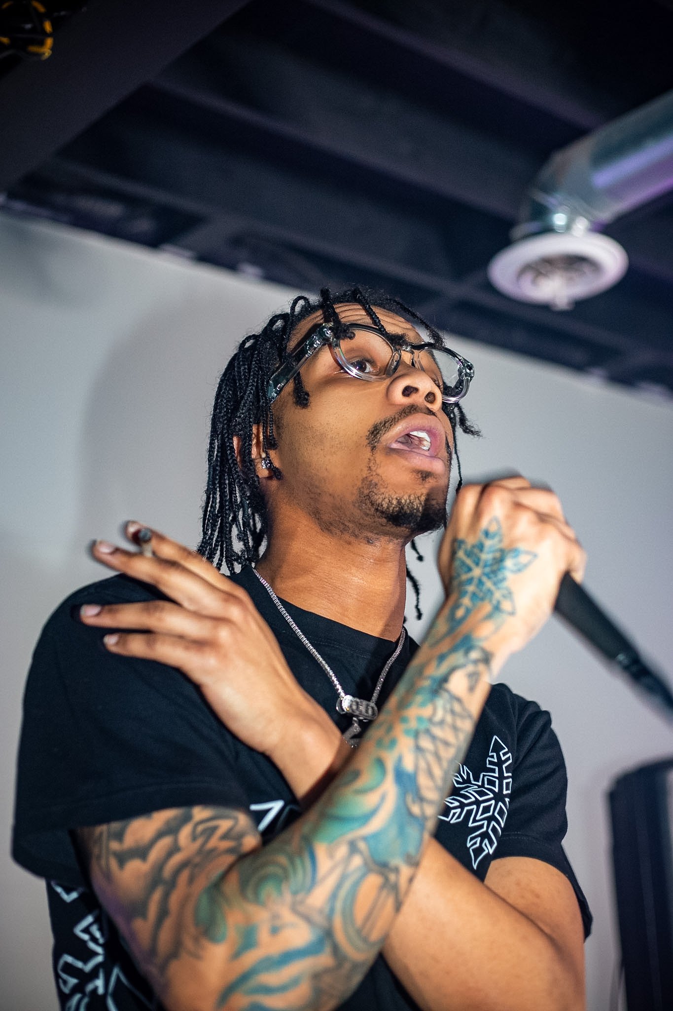 A young man with tattoos on his arm, wearing glasses, a black t-shirt, and a silver chain, holding a microphone, singing or speaking passionately with one arm crossed over his chest.