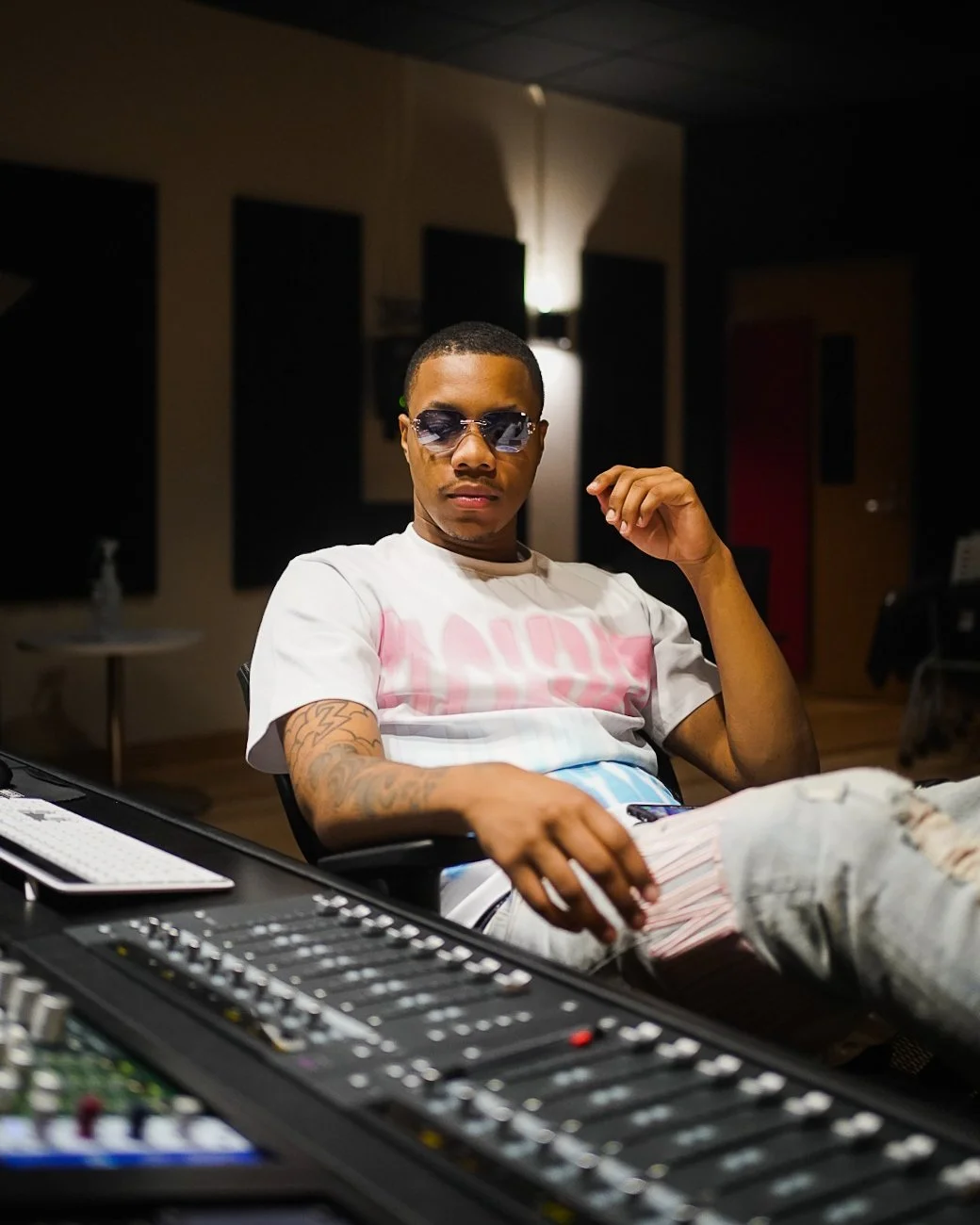 A young man wearing sunglasses, a white T-shirt with pink lettering, and ripped jeans sitting in a recording studio with audio equipment.