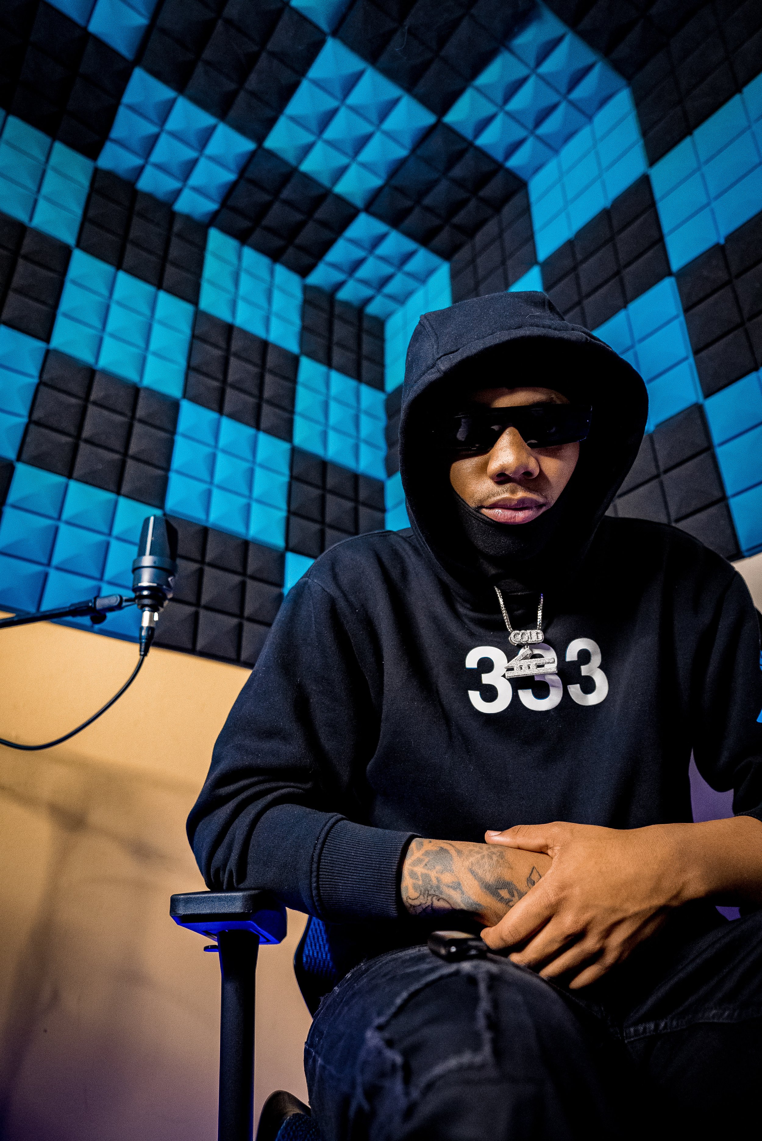 A person wearing a black hoodie, sunglasses, and a face mask sitting in a recording studio with blue and black acoustic foam panels.
