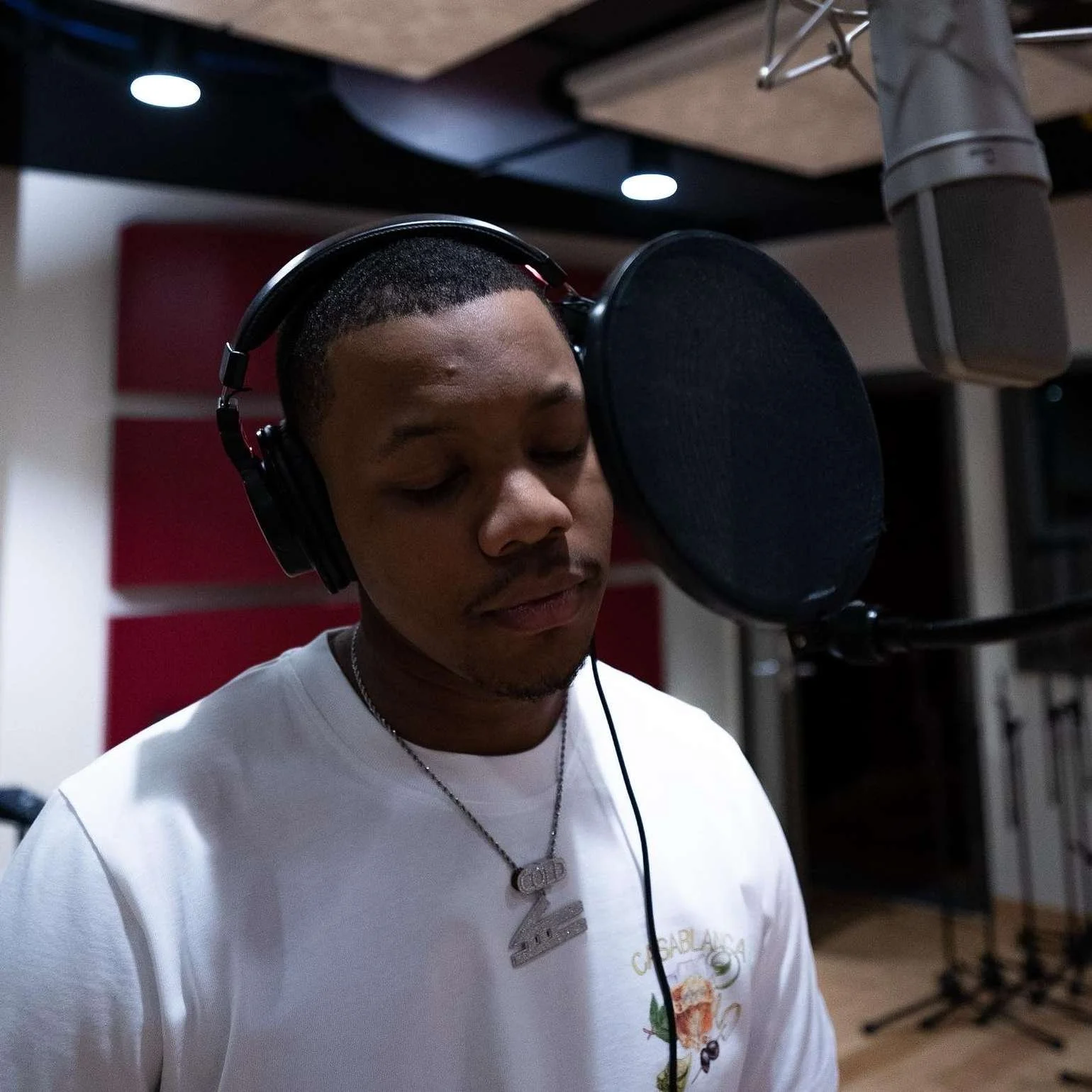 A young man with short hair and a mustache is singing or recording in a microphone in a music studio, wearing headphones and a white t-shirt with a necklace.