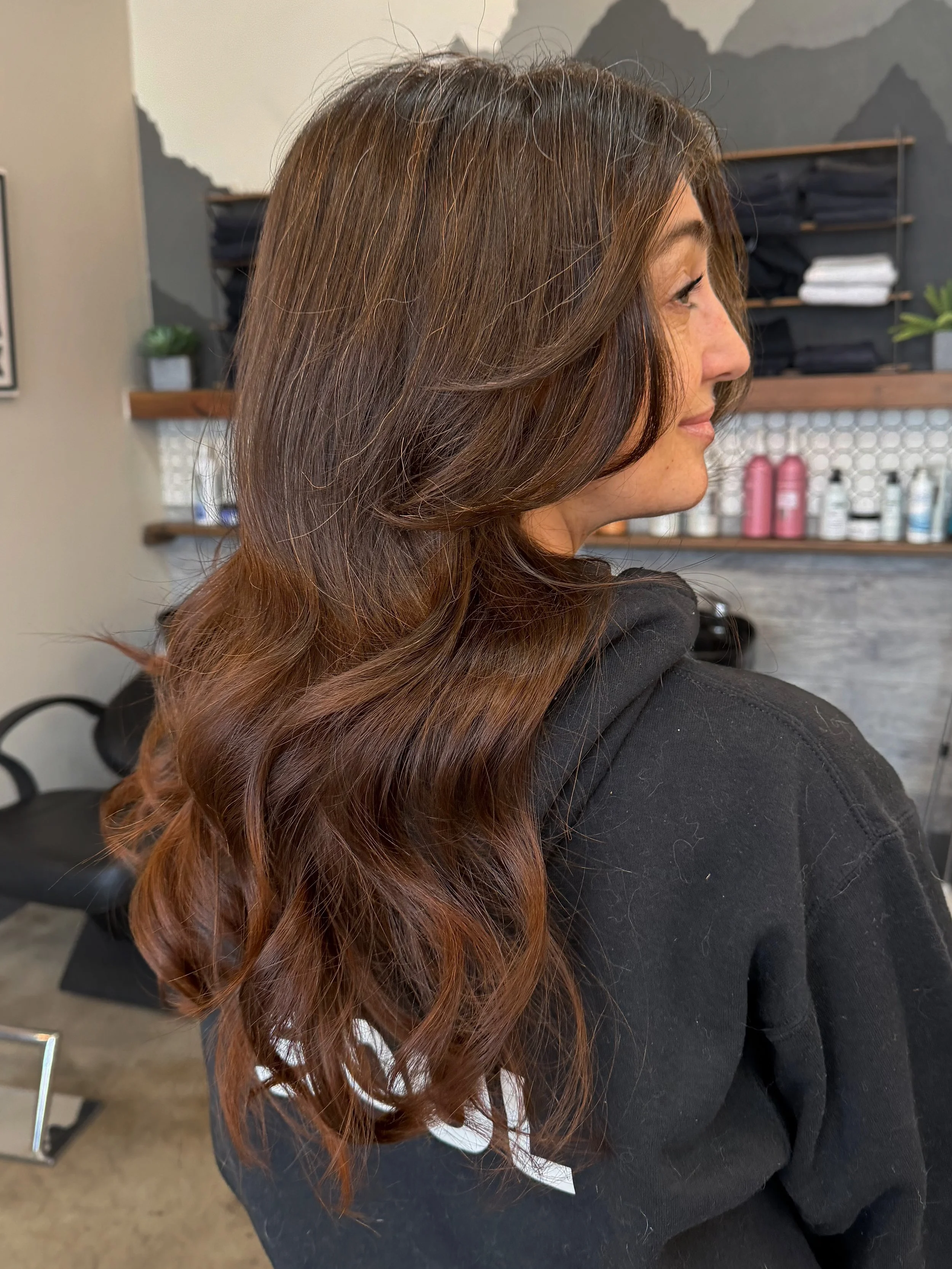 A woman with long, wavy brown hair with reddish highlights, sitting in a salon. Shelves with black and white towels, and various bottles are visible behind her.
