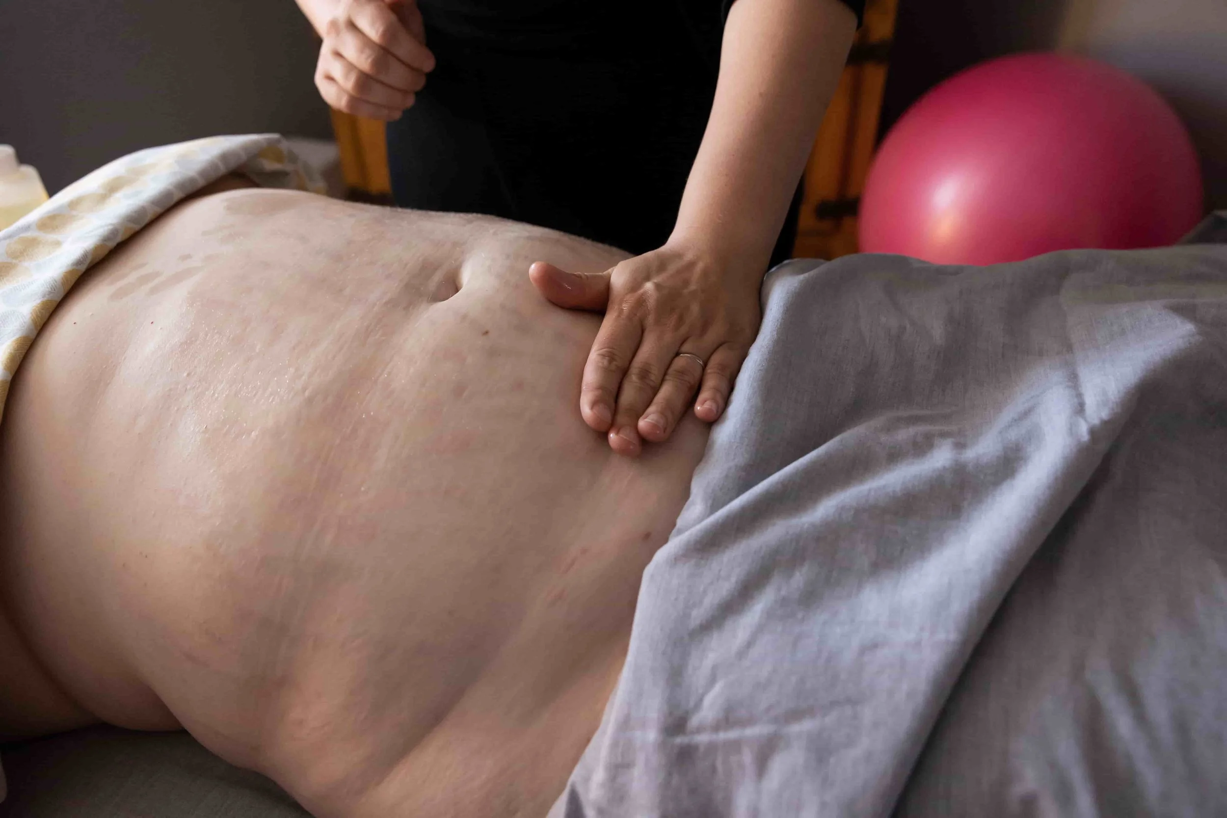 Close-up of a person lying down, receiving a massage on their bare abdomen, with a pink exercise ball in the background.