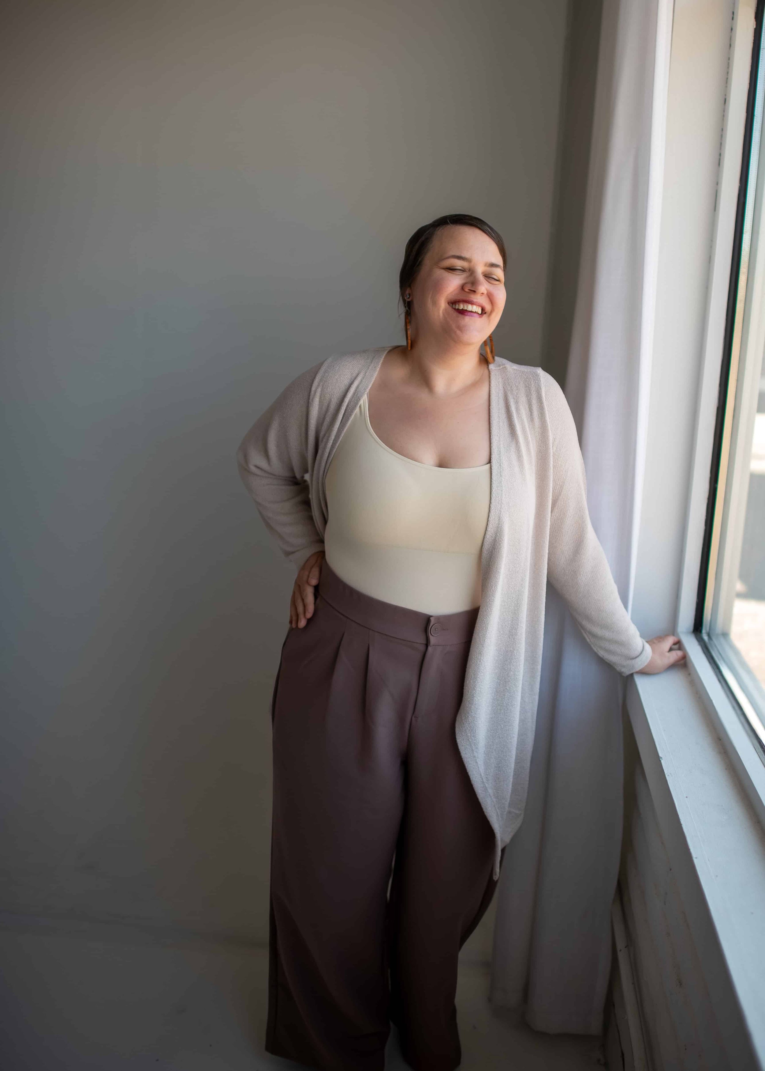 Smiling woman with short brown hair, wearing a light beige tank top, beige cardigan, and high-waisted brown pants, standing by a window with her eyes closed in a bright room.