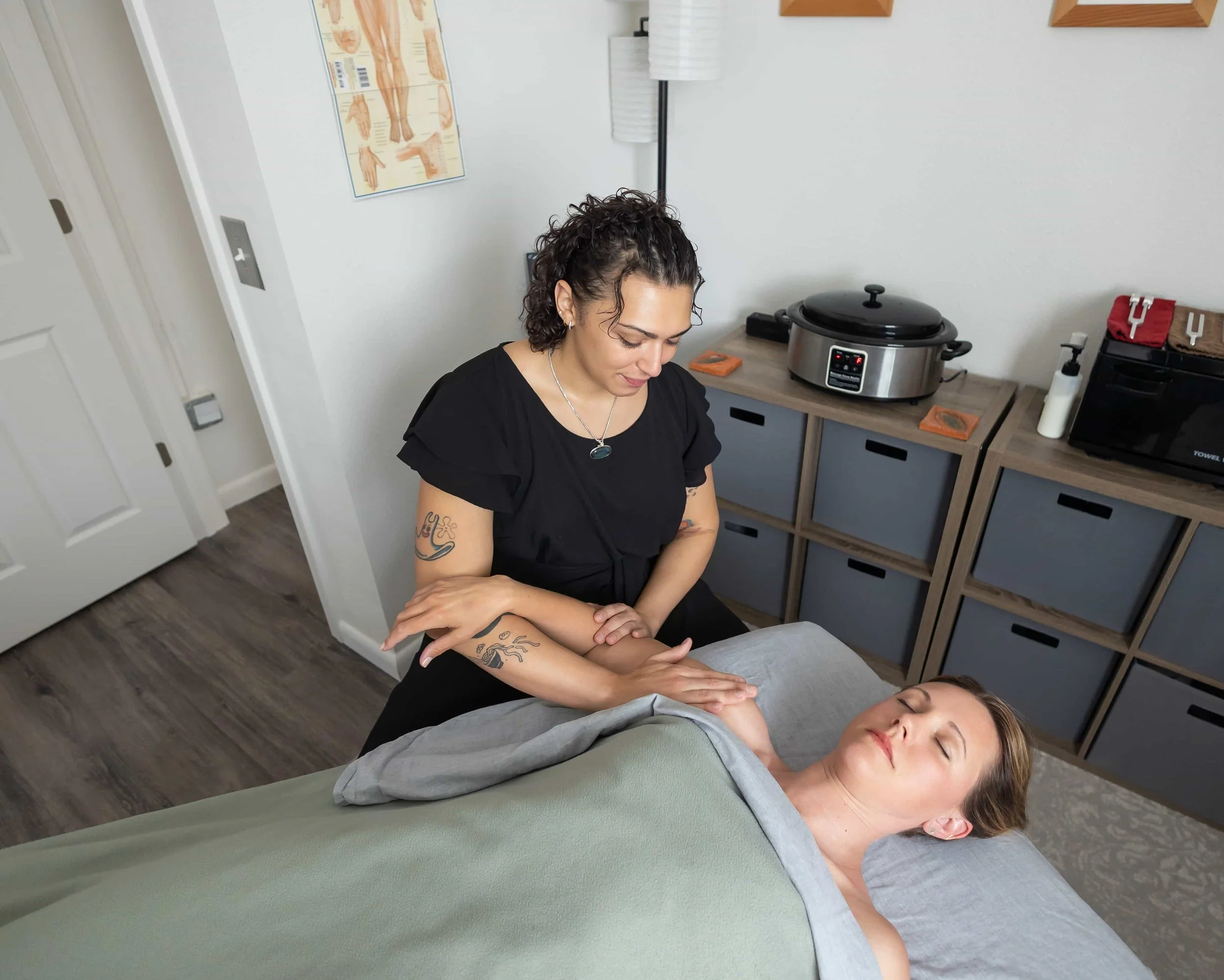 A woman with curly hair giving a massage to a woman with straight hair who is lying down with eyes closed, in a treatment room.