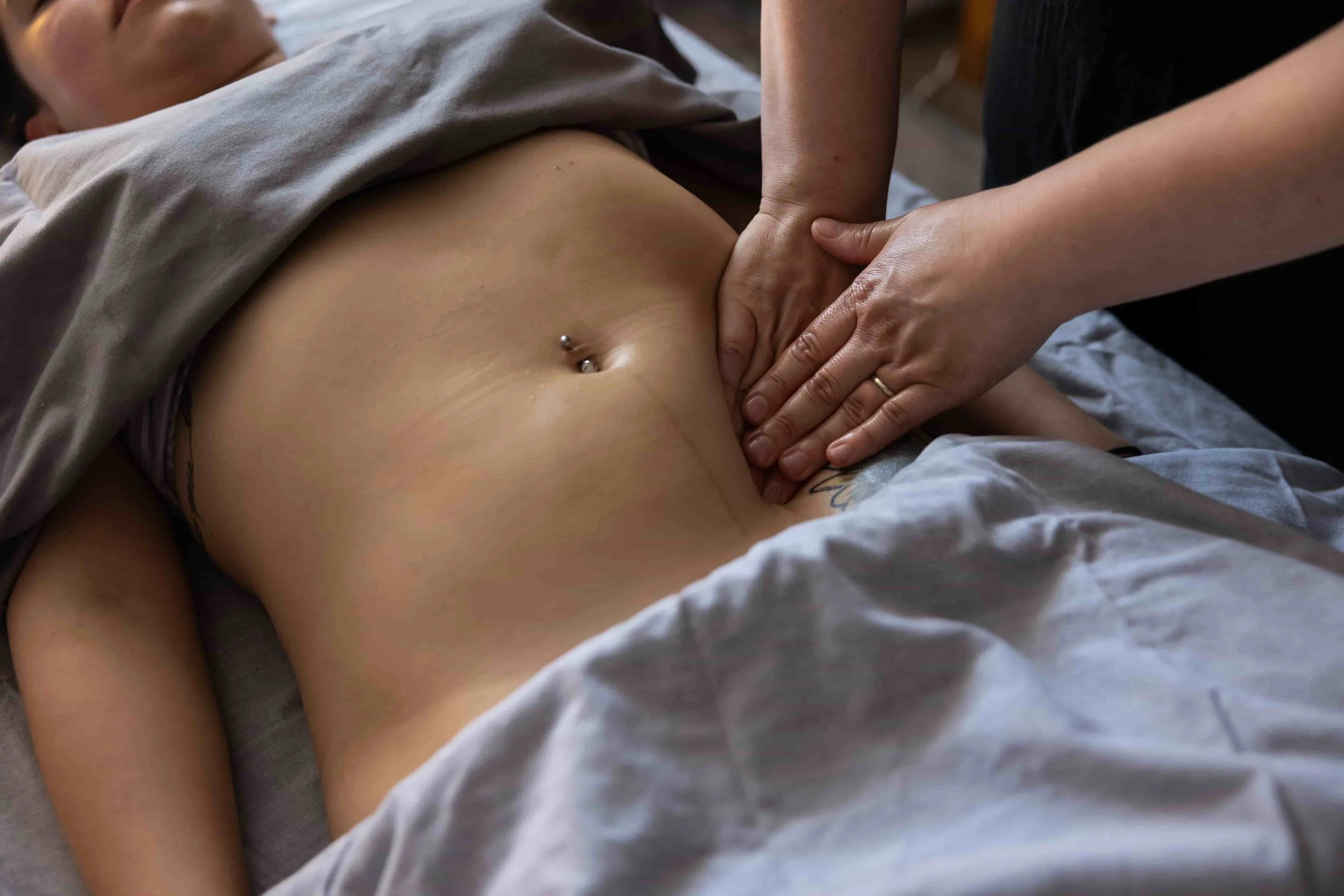 Close-up of a person's bare midsection with a belly button piercing, lying on a bed covered with a gray blanket, with hands pressing on their stomach.