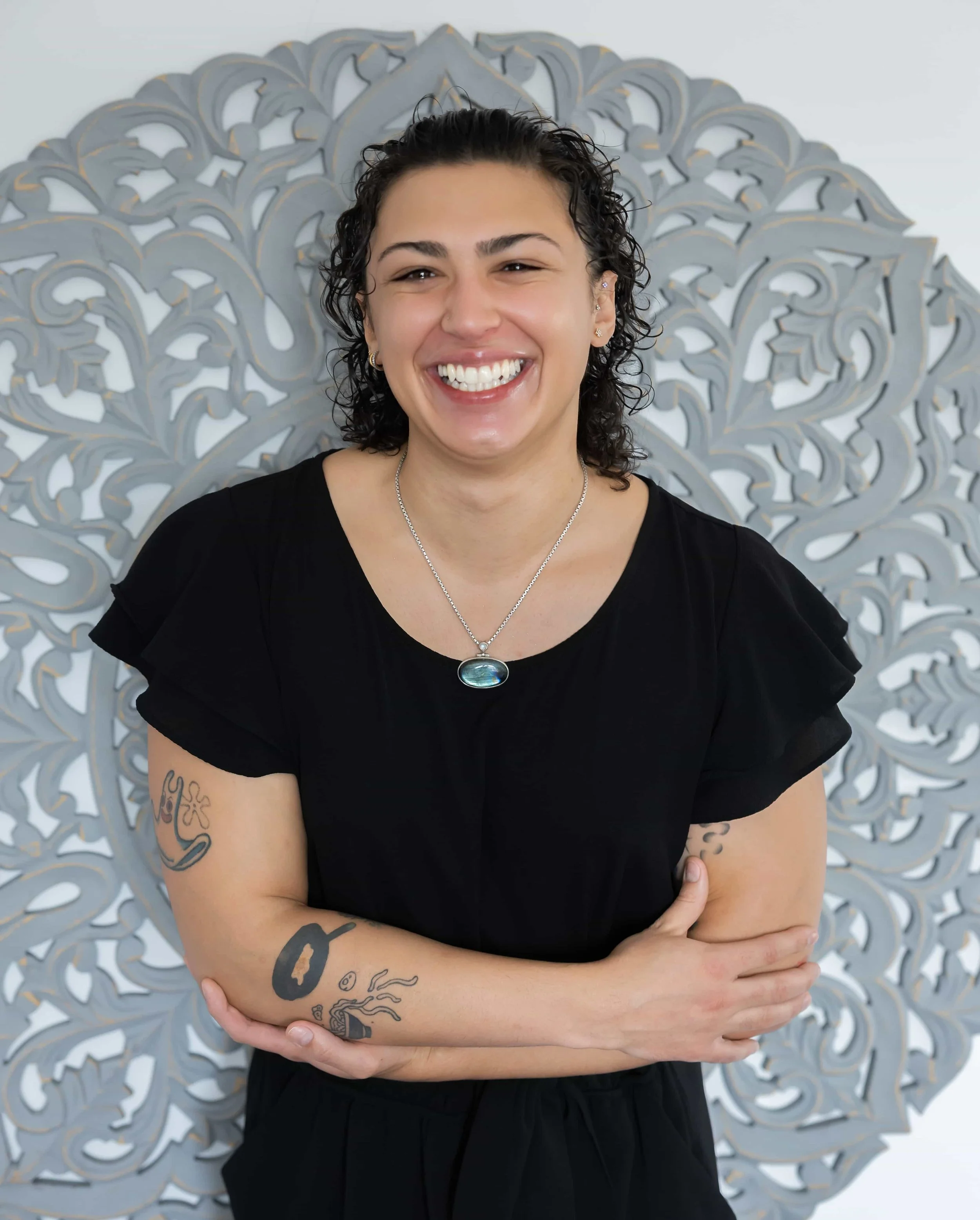 A woman with short curly dark hair smiling, wearing a black top, silver necklace, and earrings, standing against a decorative silver-gray background.