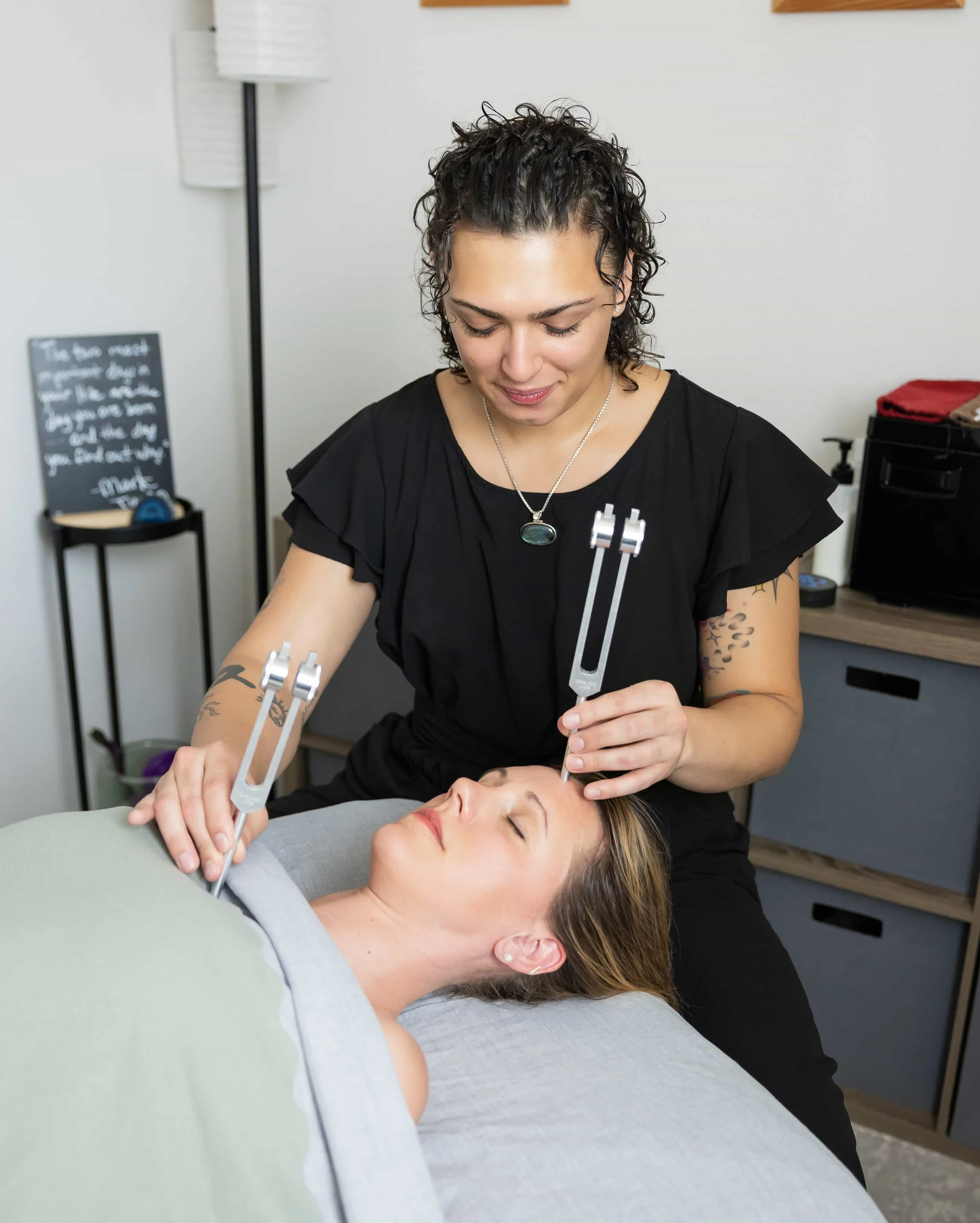 A woman is lying on a treatment bed with her eyes closed, while another woman is performing a facial treatment using tools on her face in a spa or clinic setting.
