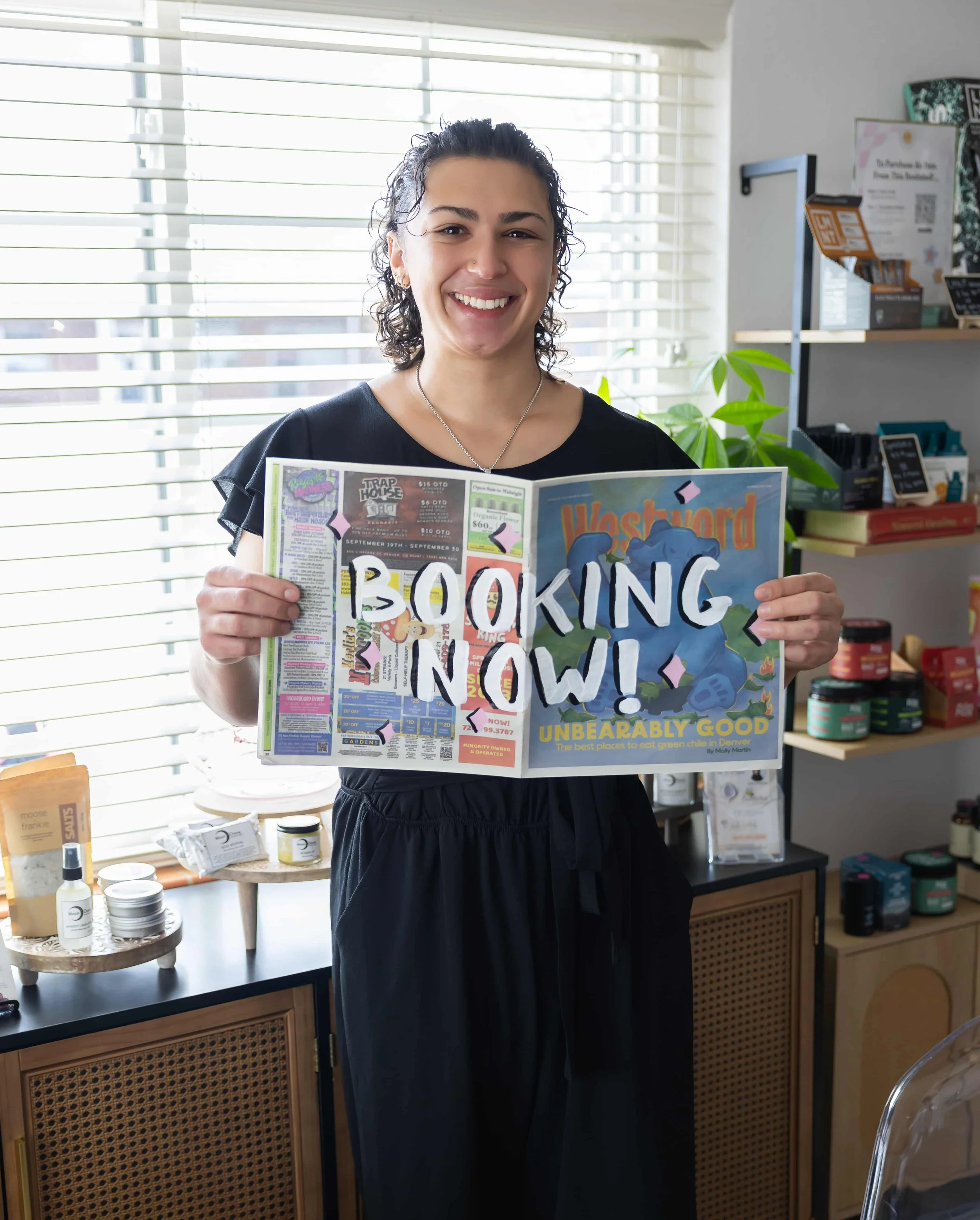 A woman smiling and holding a colorful magazine inside a retail shop with various products on shelves and a window with blinds behind her.
