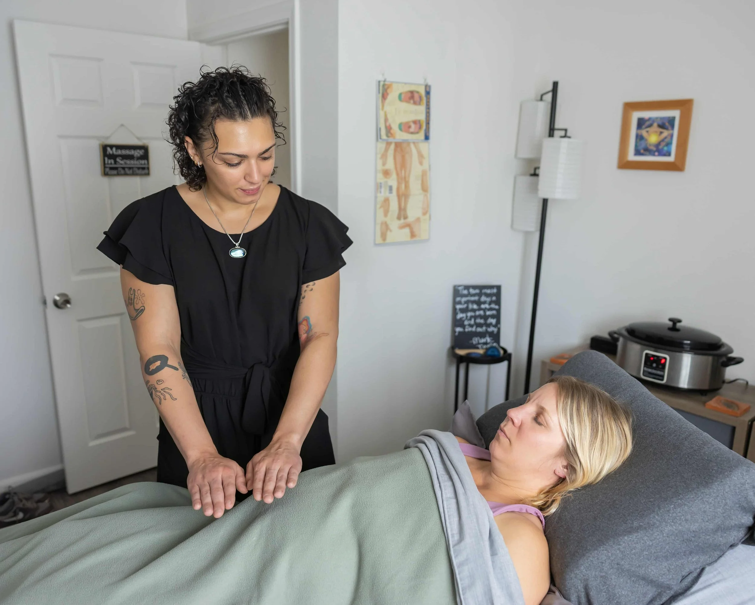 A woman lying on a treatment table receiving a therapy, with a therapist standing beside her holding her arms. The therapist has tattoos on her arms and is looking down, while the woman appears relaxed with her eyes closed. The room has medical decorations, including a visual chart of the human body, and art on the walls. There is a black chalkboard with writing, a standing lamp, and medical equipment in the background.