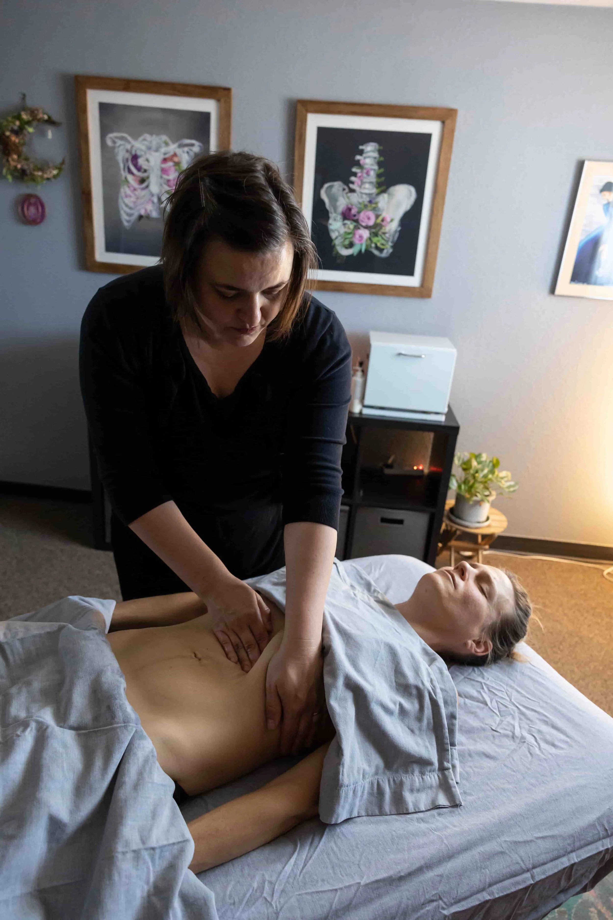 A woman receiving a massage from a massage therapist on a massage table in a cozy room with framed art on the walls and a small table with a plant.