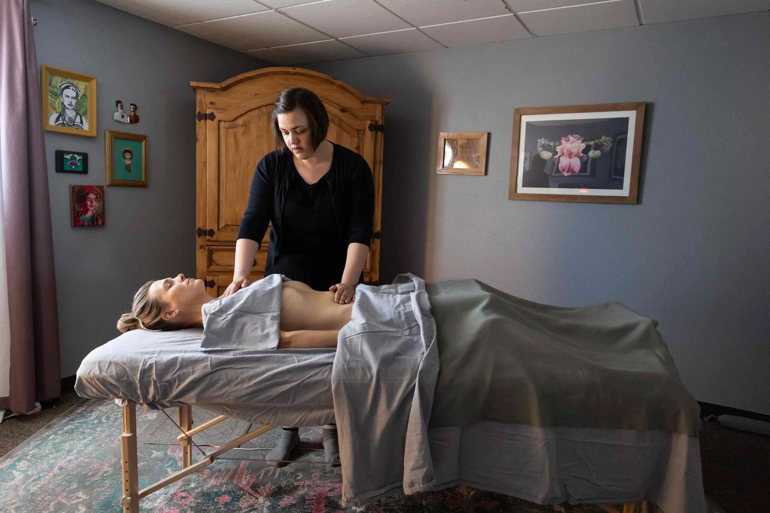 A woman receiving a massage on her back while lying on a massage table, with a massage therapist standing beside her, in a room decorated with framed artwork on the wall.