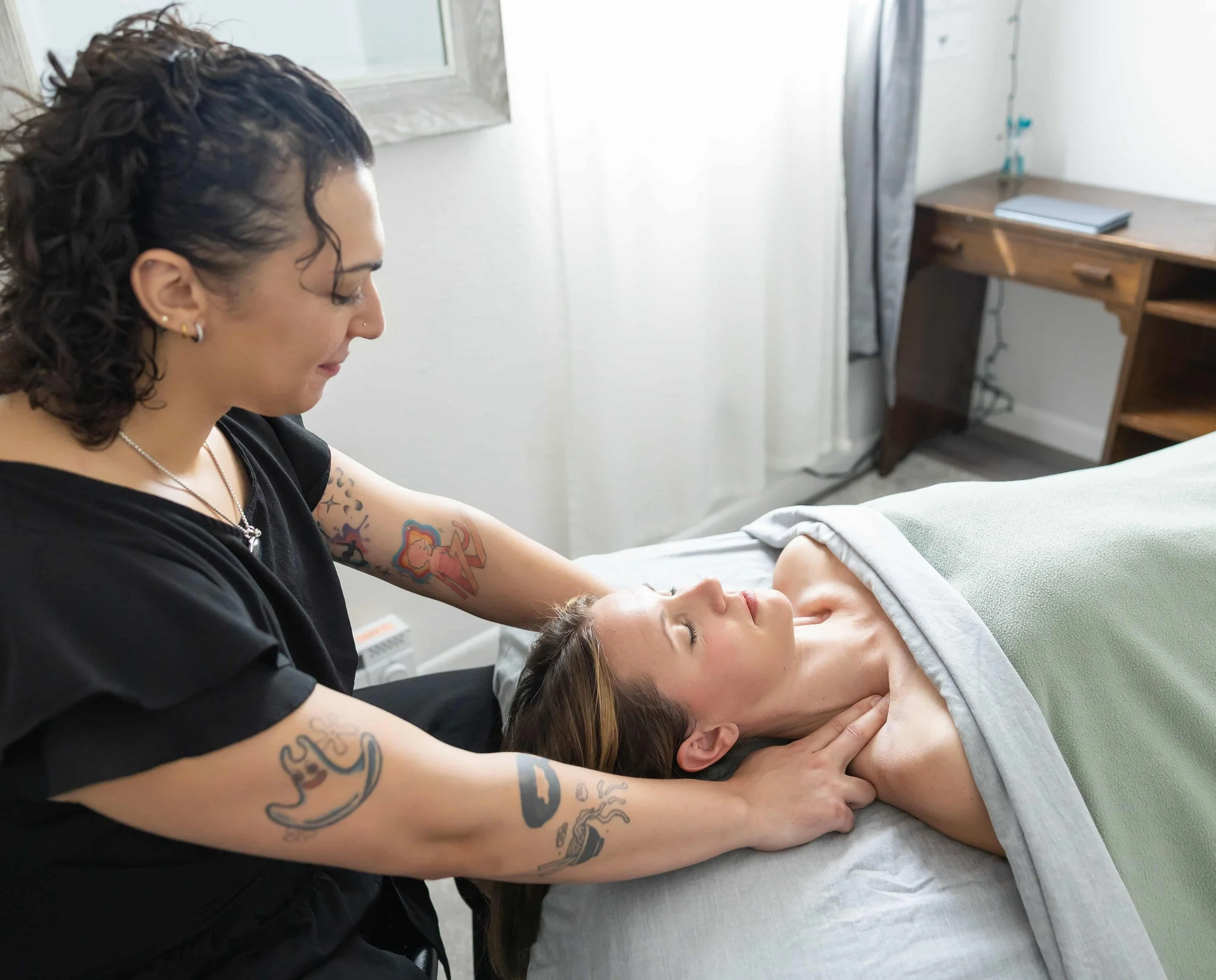 A woman giving a massage to a woman lying on a massage table in a room with white walls and a wooden desk.
