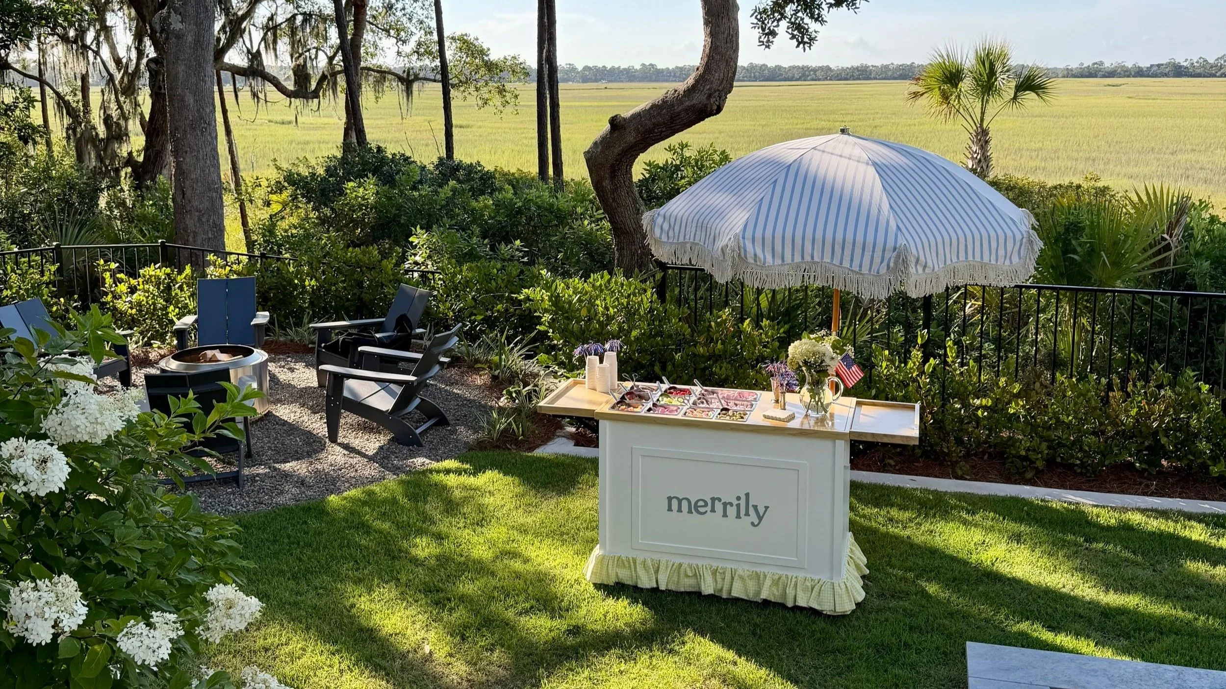 Outdoor patio with chairs, a table with snacks, a white umbrella, and lush greenery overlooking a field.