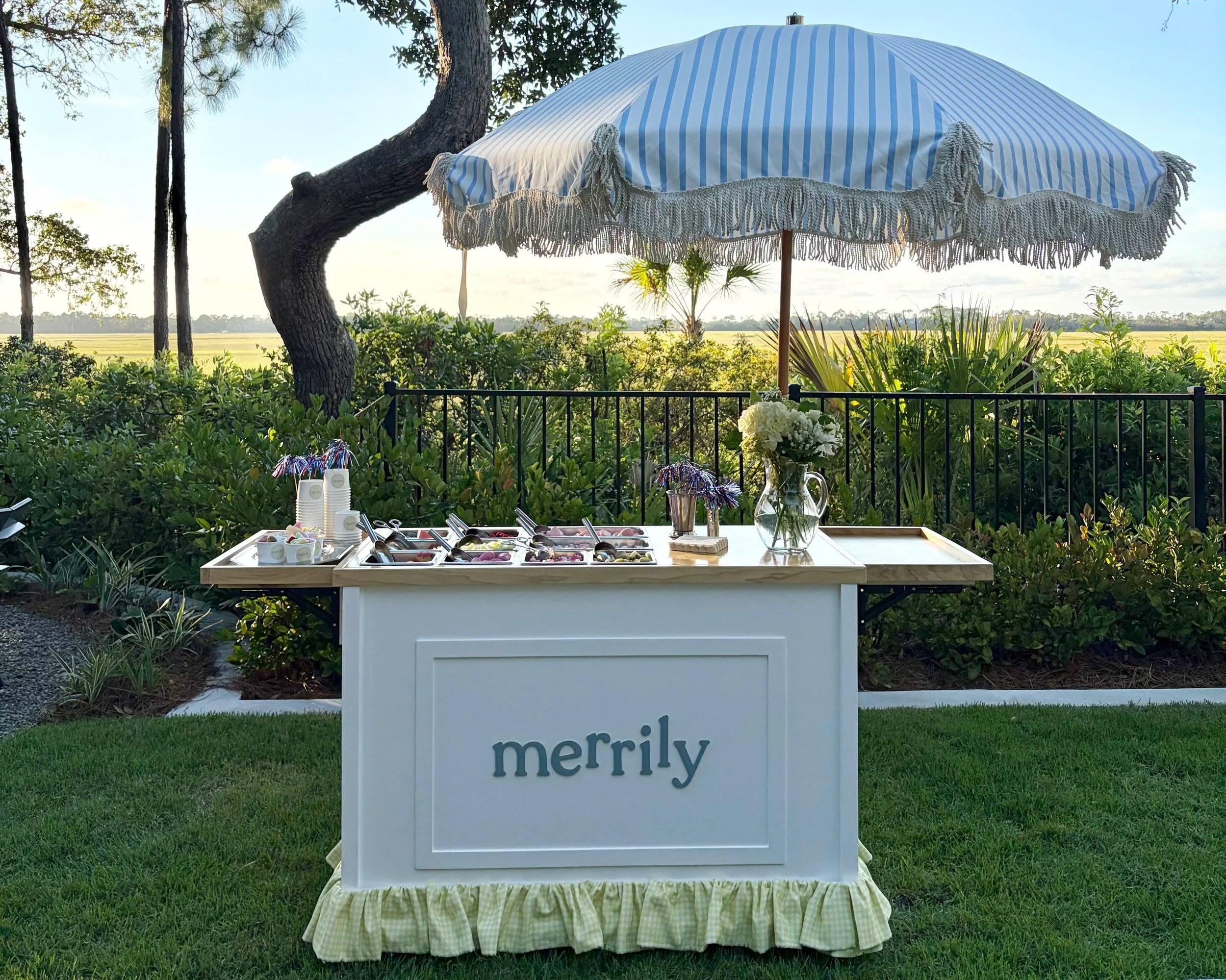 A white candy cart with a sign reading 'merrily', set outdoors with an umbrella, surrounded by bushes and trees. The table has various sweets, a yellow gingham skirt along the bottom, and flowers.