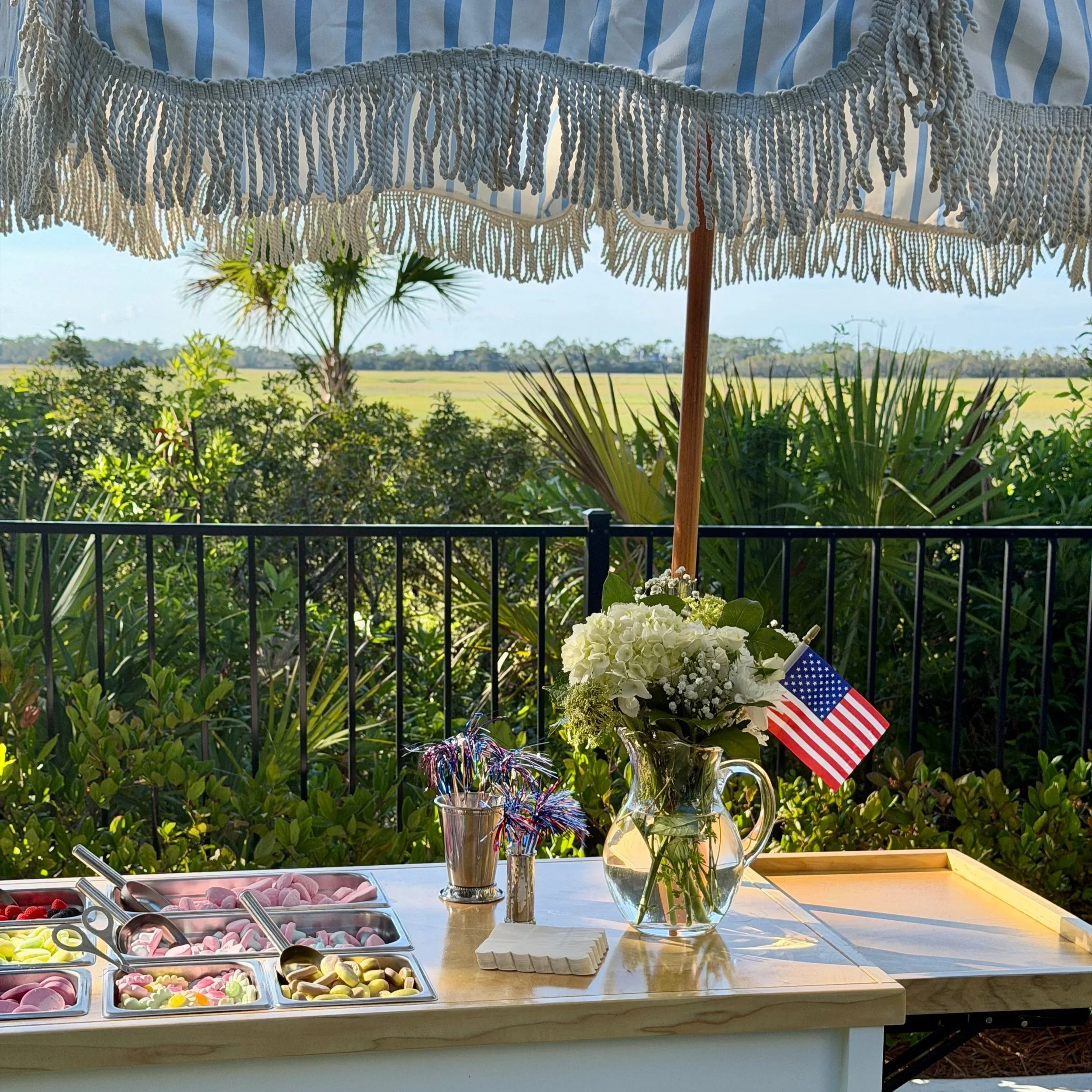 Table with assorted candies, a bouquet of white flowers, an American flag, and a silver trophy in an outdoor setting under a patio umbrella with a scenic background of fields and trees.