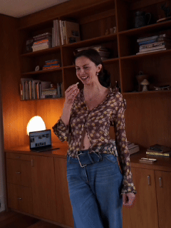 A woman smiling and standing in front of wooden shelves filled with books and decorative items in a cozy room.