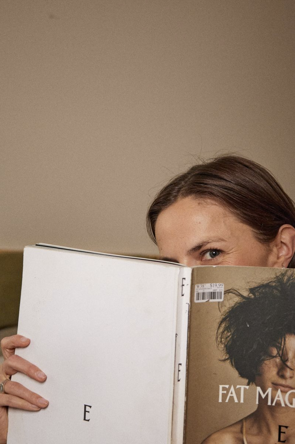 Woman with brown hair and blue eyes peeking over a magazine cover with short curly black hair, in front of a beige background.