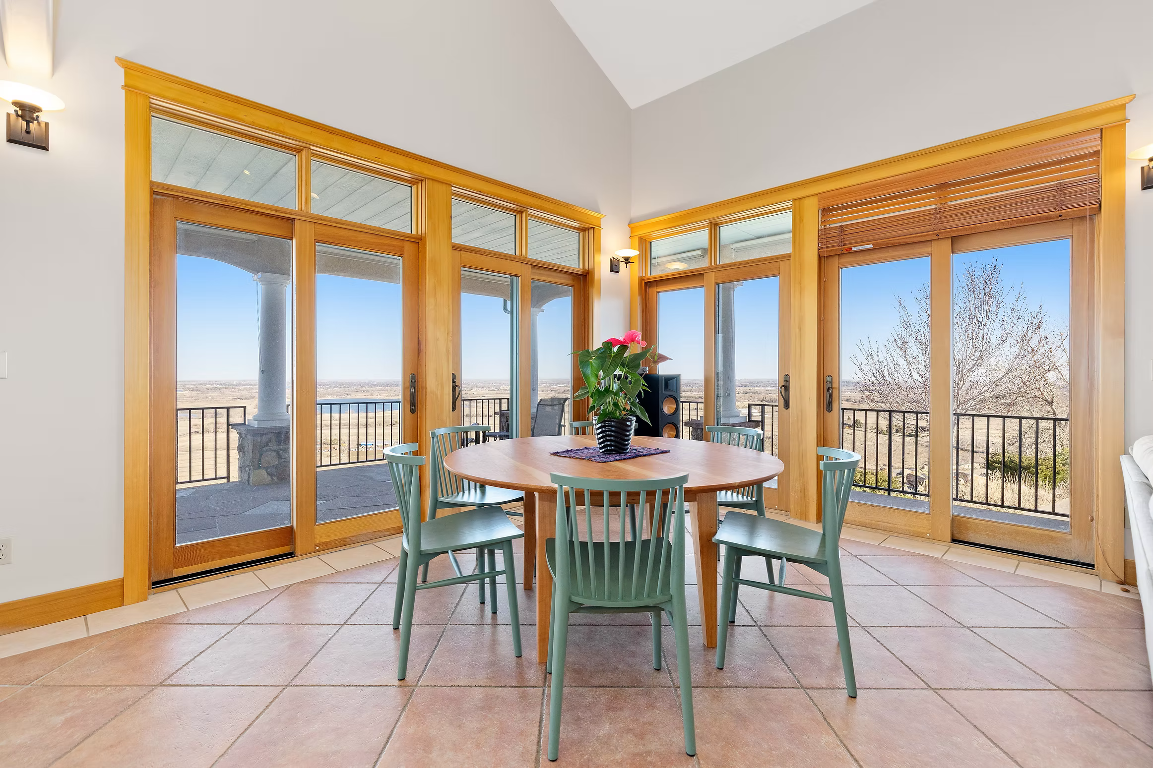 A dining room with large glass sliding doors showing an outdoor balcony with a scenic view of open land. There is a round wooden table with six teal chairs and a flowerpot with pink flowers in the center.