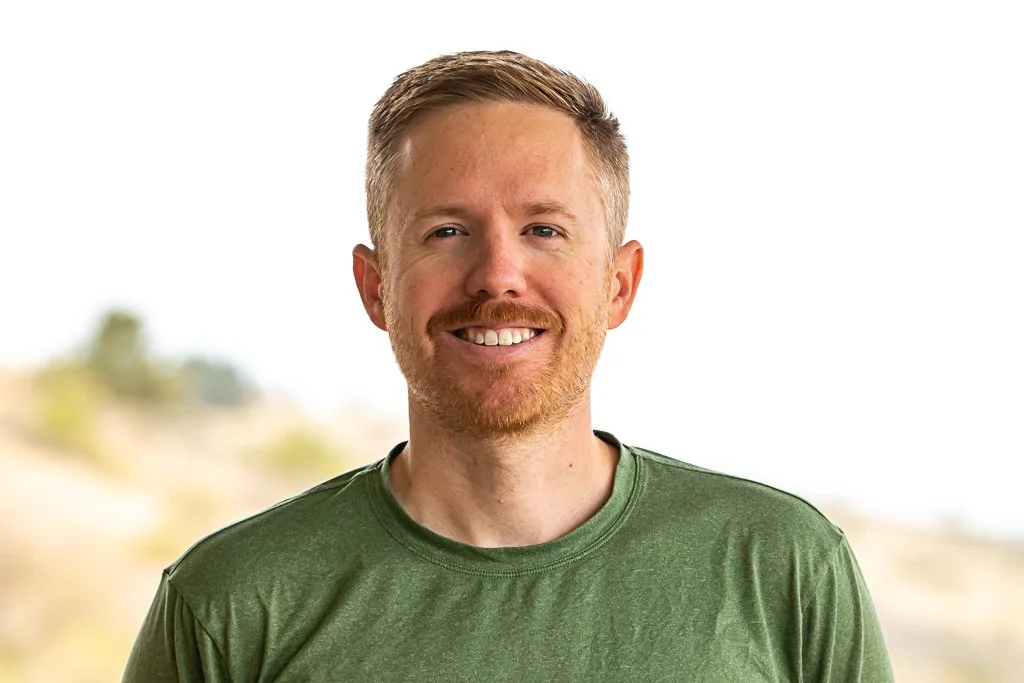 Portrait of a smiling man with short, light brown hair and a beard, wearing a green t-shirt, standing outdoors with a blurred natural background.