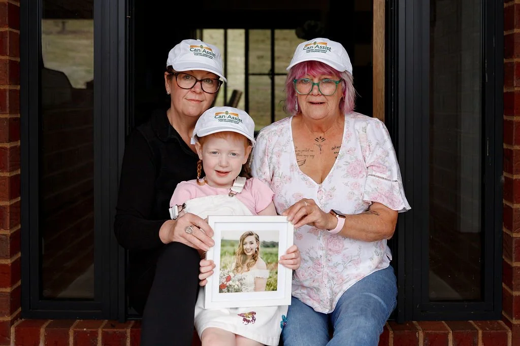 Three women, two with glasses and white caps, one young girl, sitting on a brick porch. The young girl holds a framed photo of a smiling woman outdoors in a field. The woman on the right has pink hair, tattoos on her chest, and wears a floral blouse. The woman on the left wears a black shirt. They are all looking at the camera.