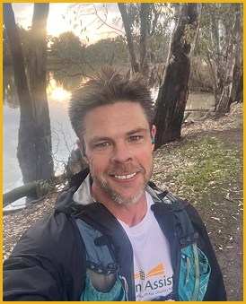 A man taking a selfie outdoors near a lake with trees, at sunset. He has short hair, a beard, and is wearing a dark jacket and a white shirt.