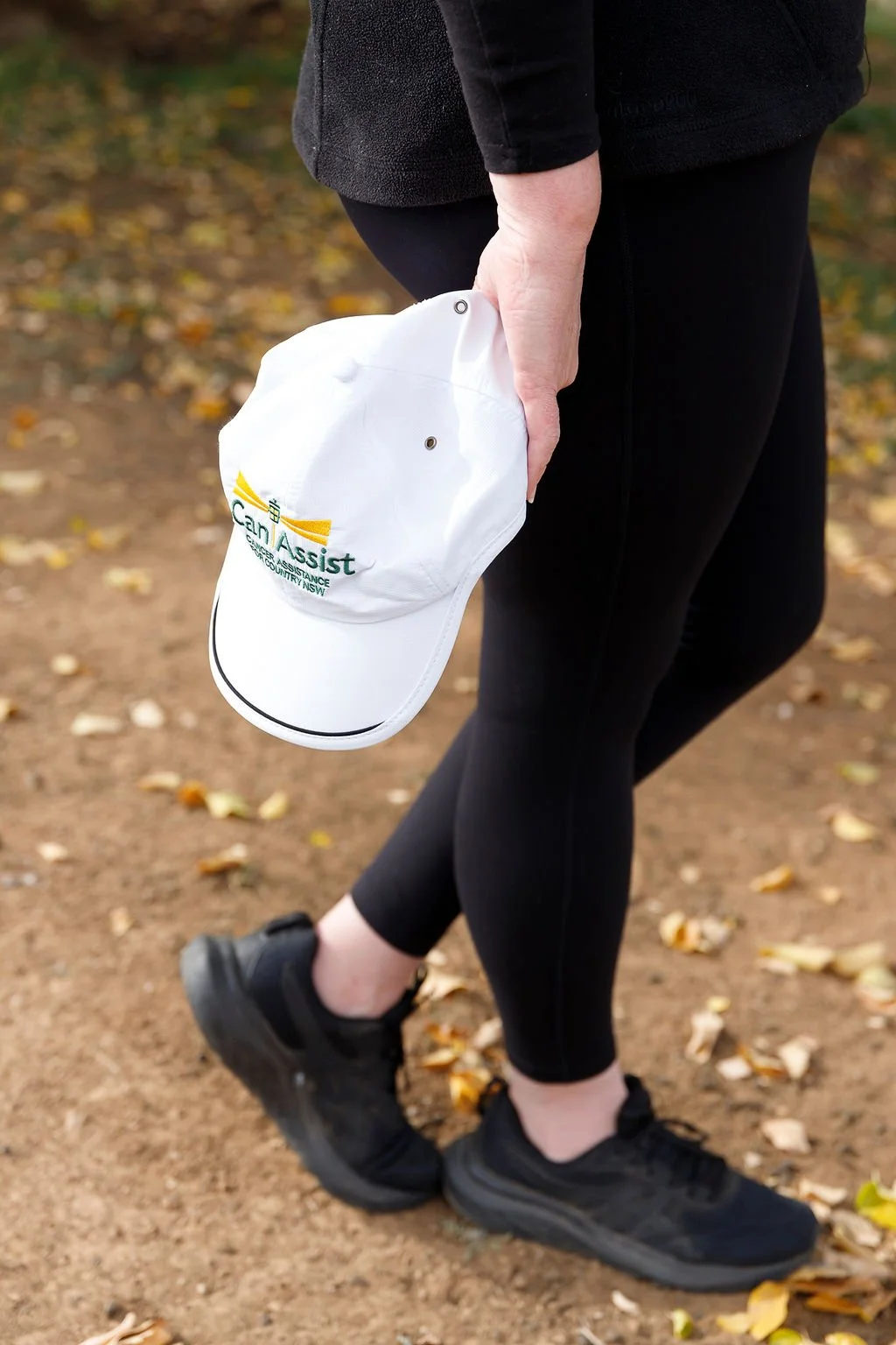 Person holding a white baseball cap with 'Can Assist' logo in their right hand, standing on a dirt path with fallen yellow leaves, wearing black athletic shoes, black leggings, and a black fleece jacket.