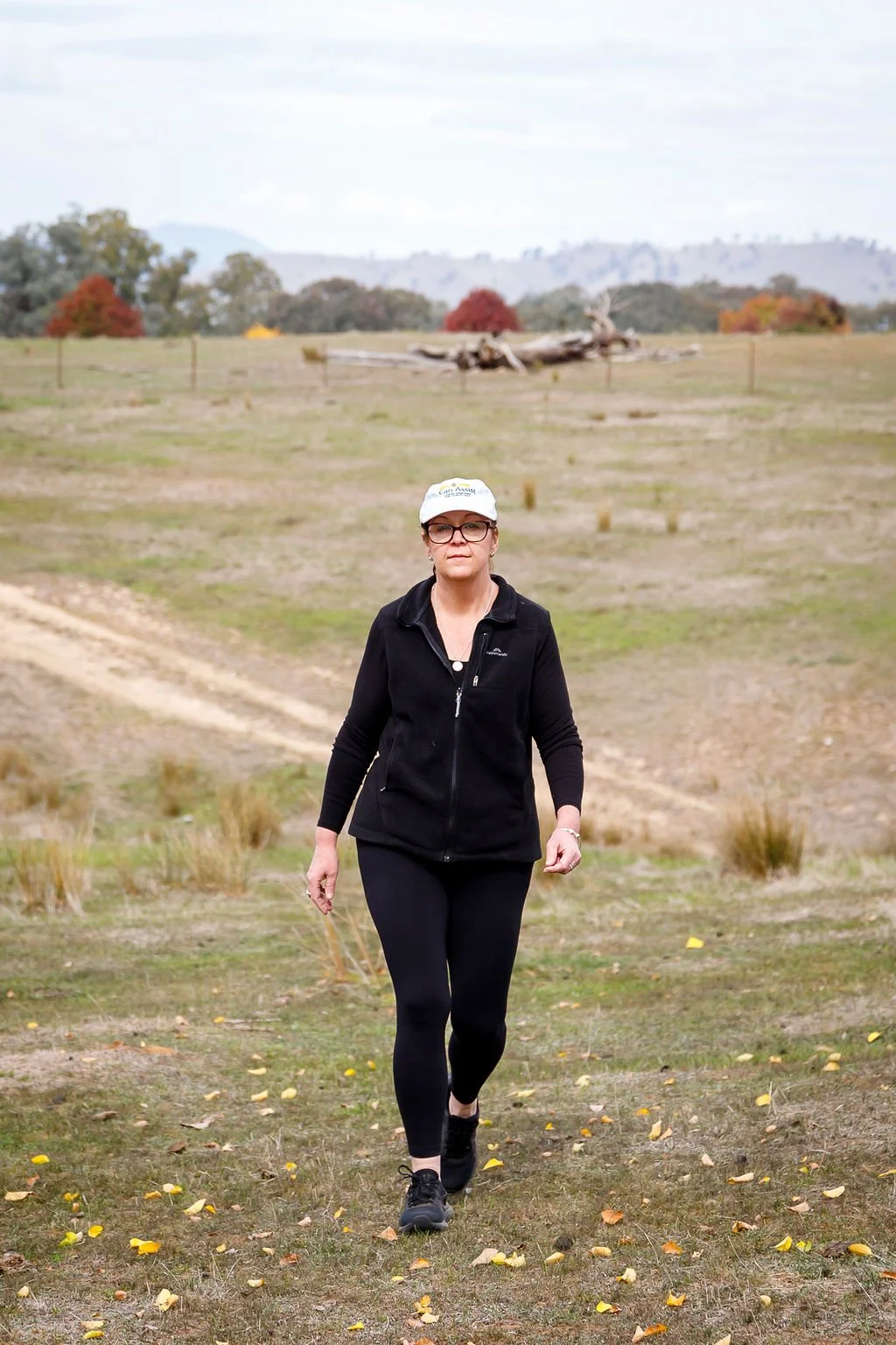 A woman wearing black athletic clothing and glasses walking on a grassy trail in an open rural landscape during daytime with trees and hills in the background.