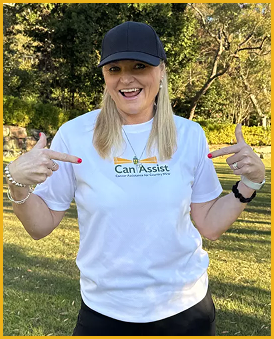Woman smiling outdoors, pointing at her shirt with both hands, wearing a navy cap and white T-shirt that says 'Can Assist.'