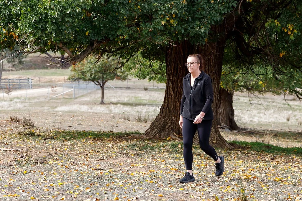 A woman walking outdoors on a dirt path covered with fallen leaves near a large tree with lush green foliage. She is wearing glasses, a black jacket, black leggings, and black shoes.